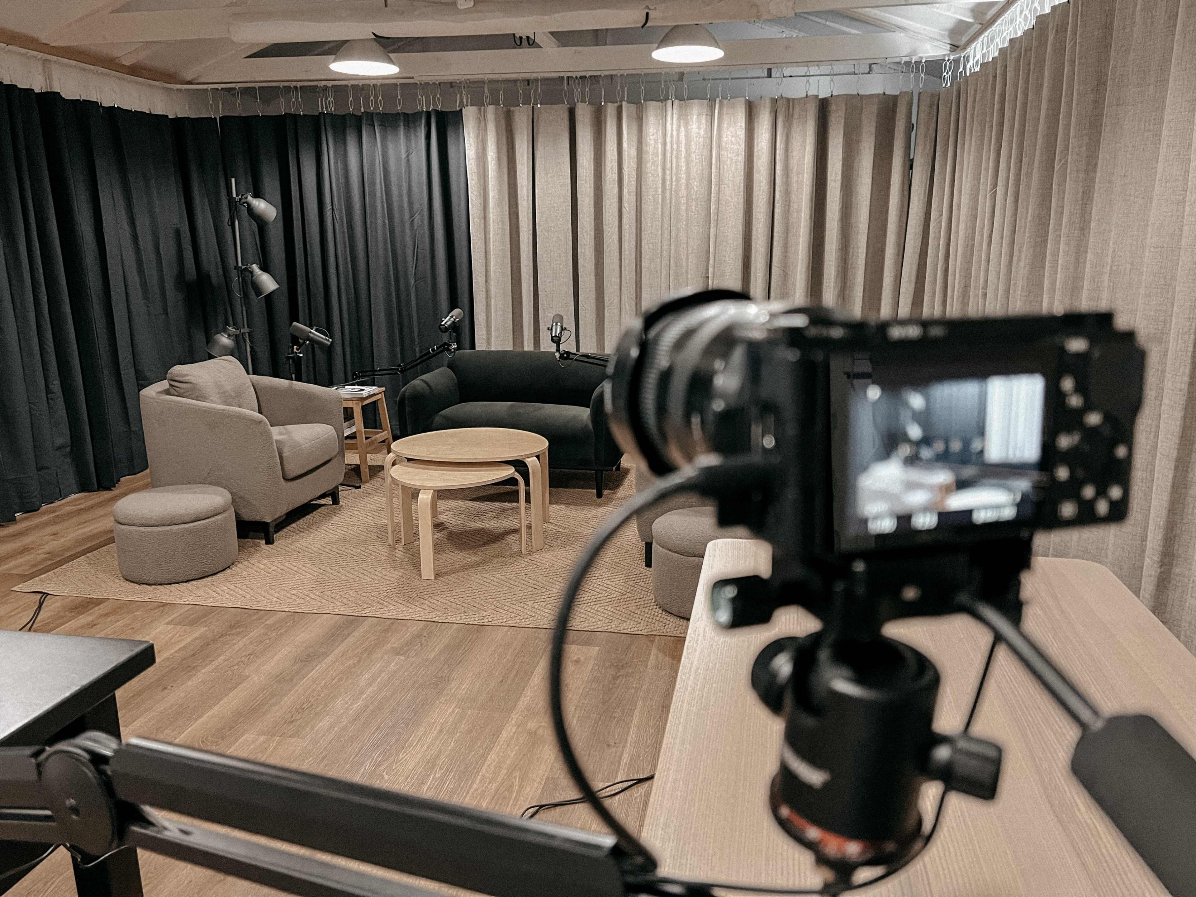 A camera focuses on a modern studio setup with beige and black furniture, including chairs and a table, surrounded by curtains.