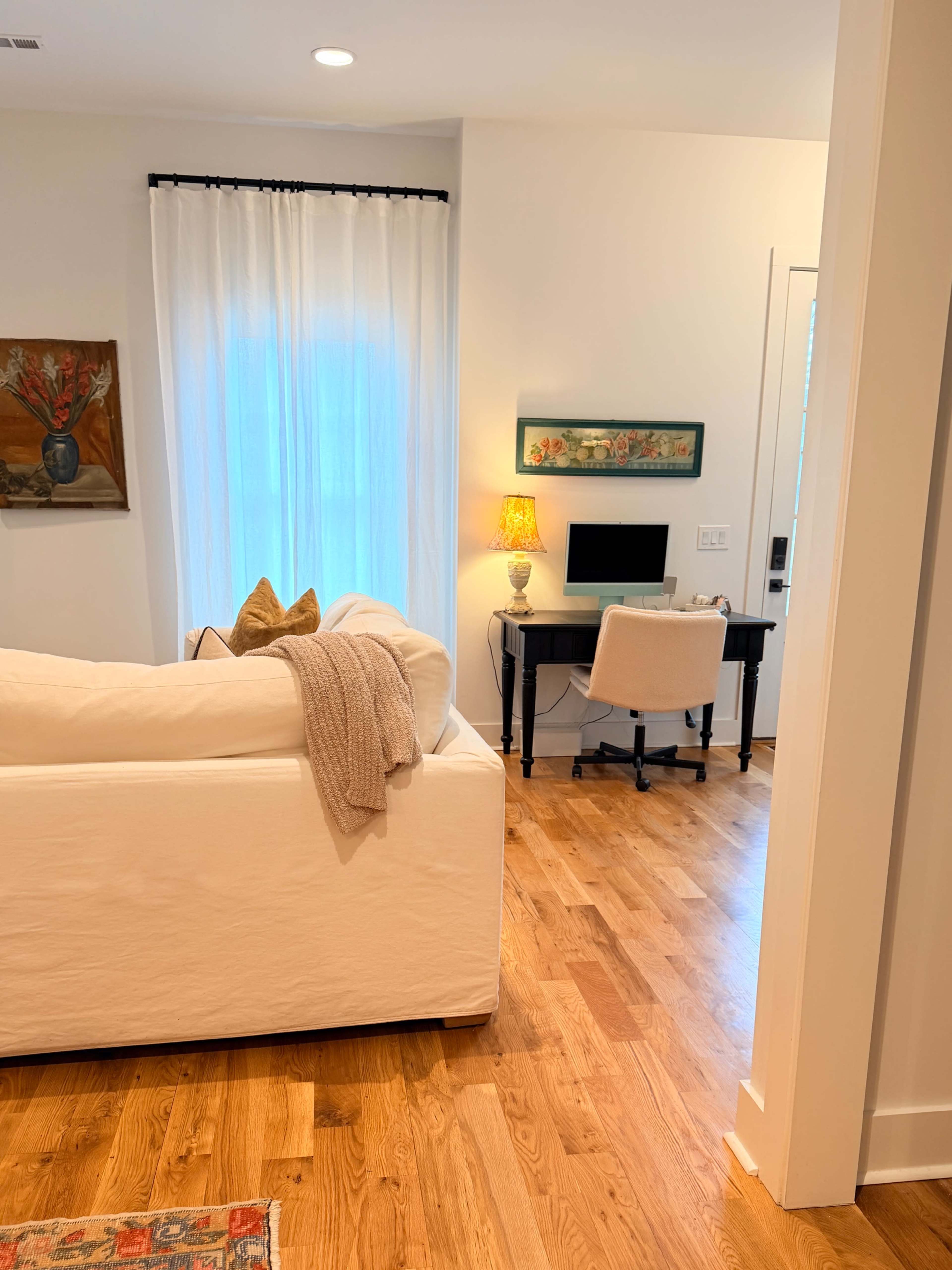 A bright room features a white sofa, a desk with a computer, and a decorative lamp, along with wooden flooring and sheer curtains.