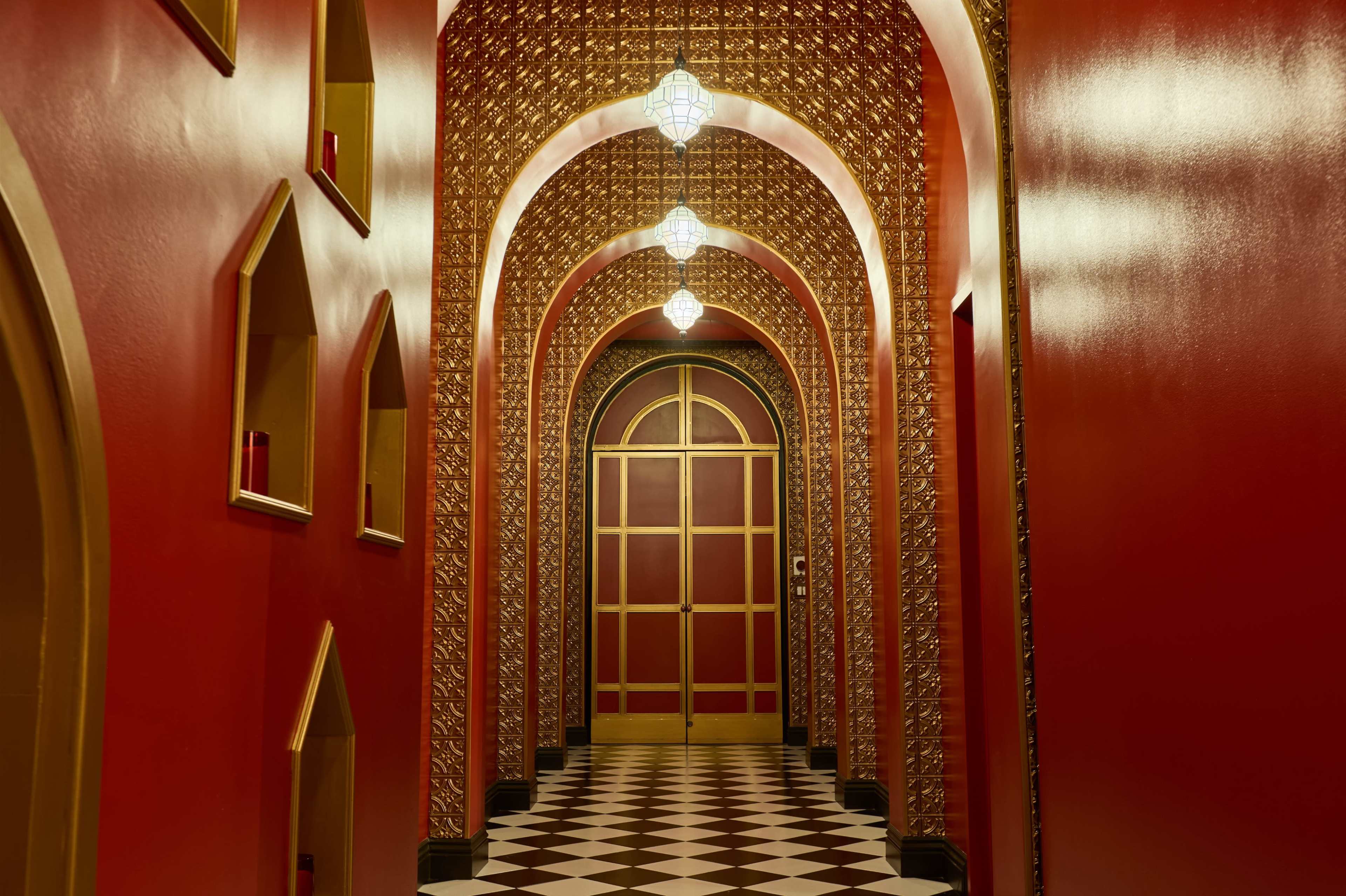 A long corridor with red walls and arches features decorative golden details and a checkered floor, leading to a large, ornate door.