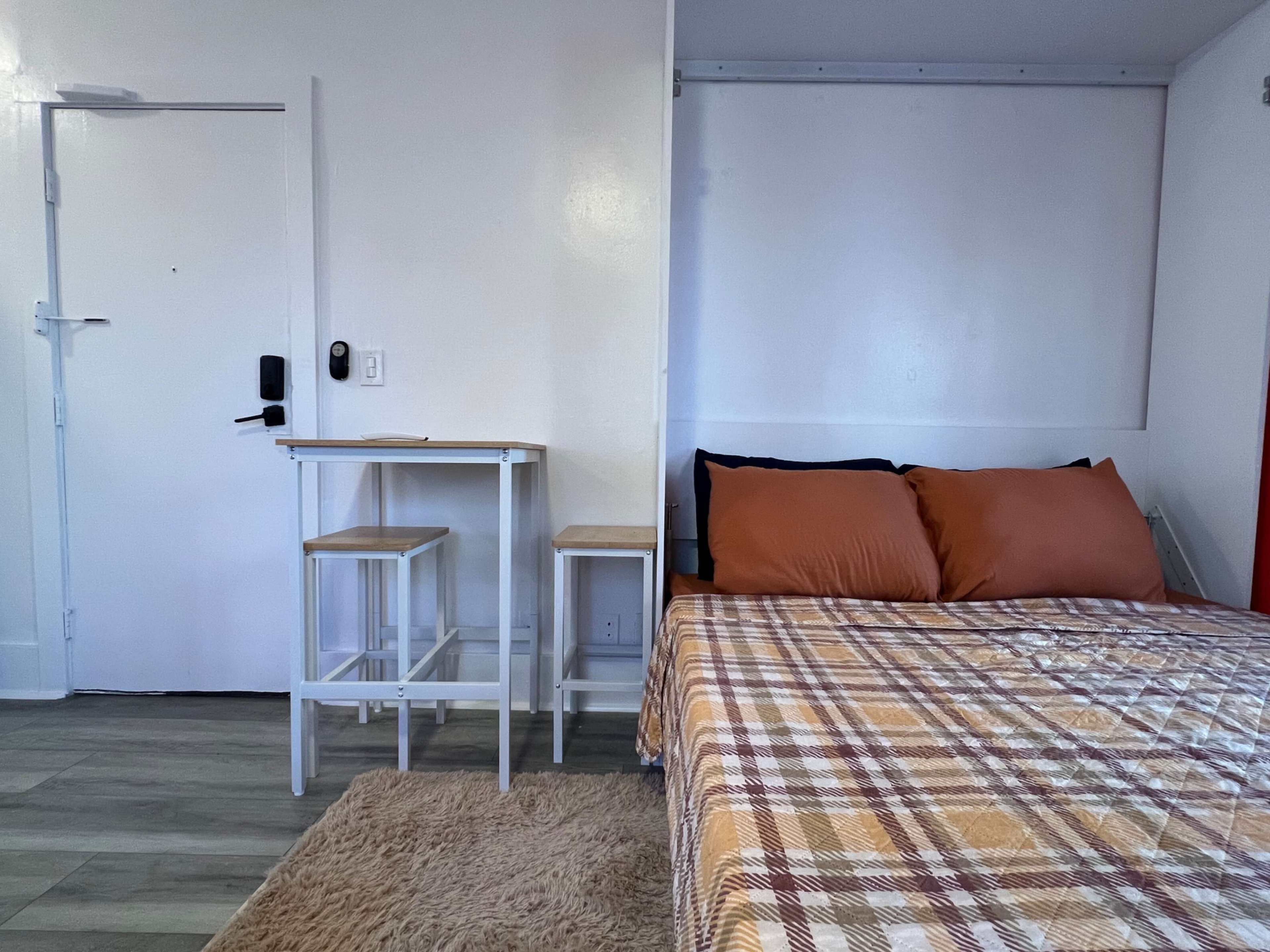 The image shows a small room featuring a bed with orange pillows, a checkered blanket, a rug on the floor, and a small table with two stools beside a door.