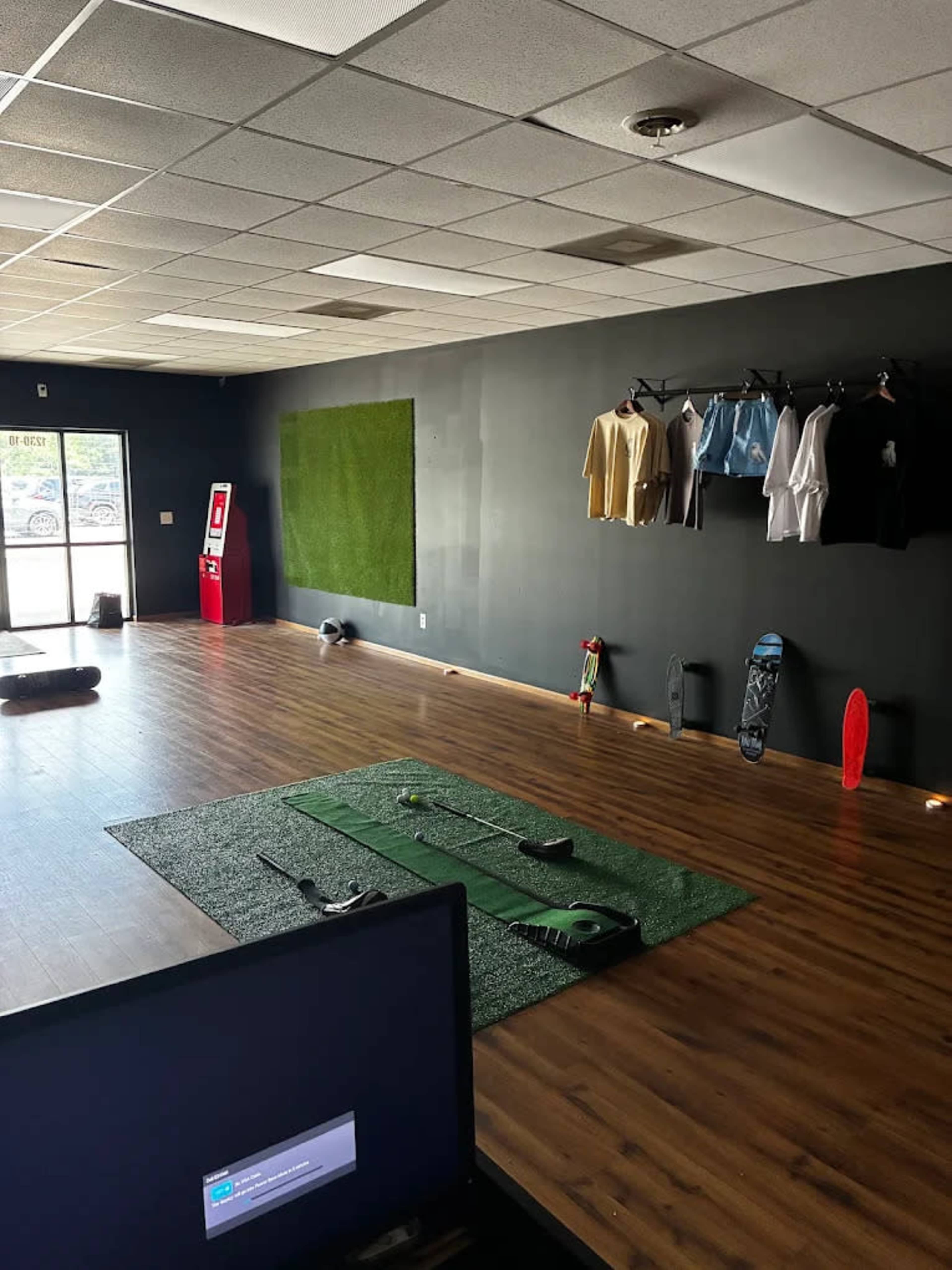 The image shows an empty indoor space with a wooden floor, a green turf patch in the center, golf clubs and equipment laid out, and several shirts hanging on a wall.