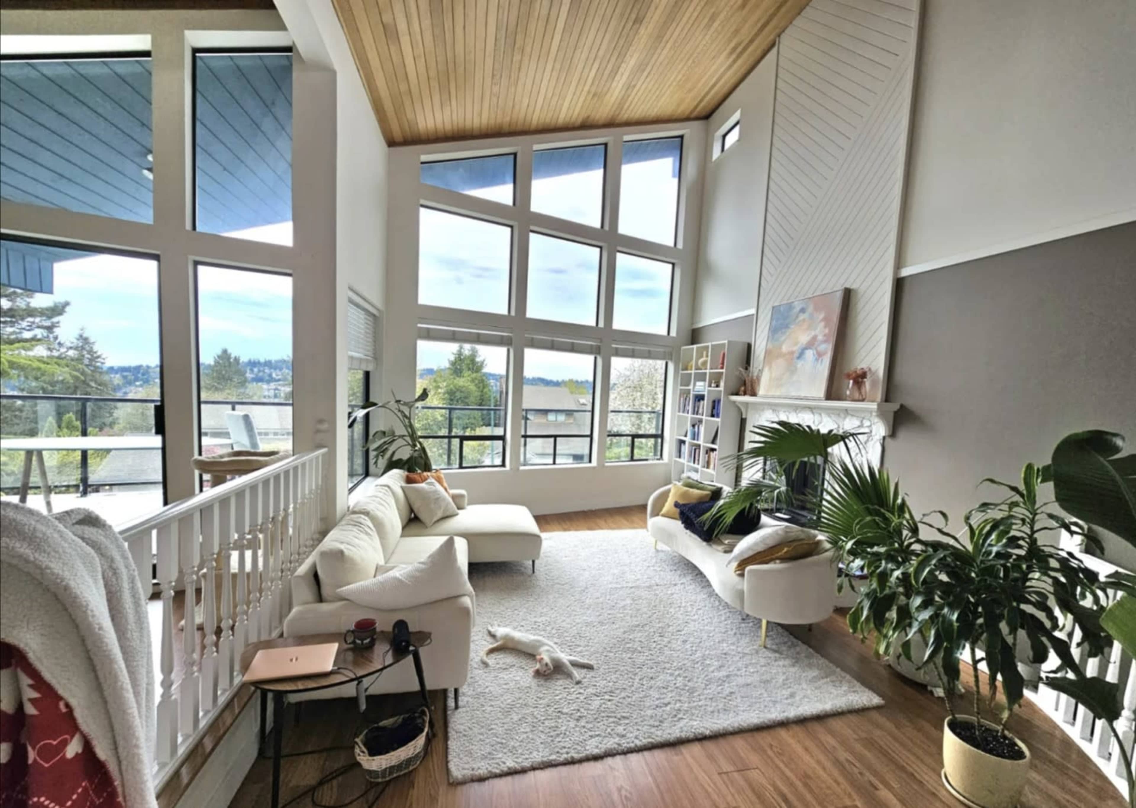 The image shows a modern living room with large windows, wood accents, and furniture arranged around a central area rug.