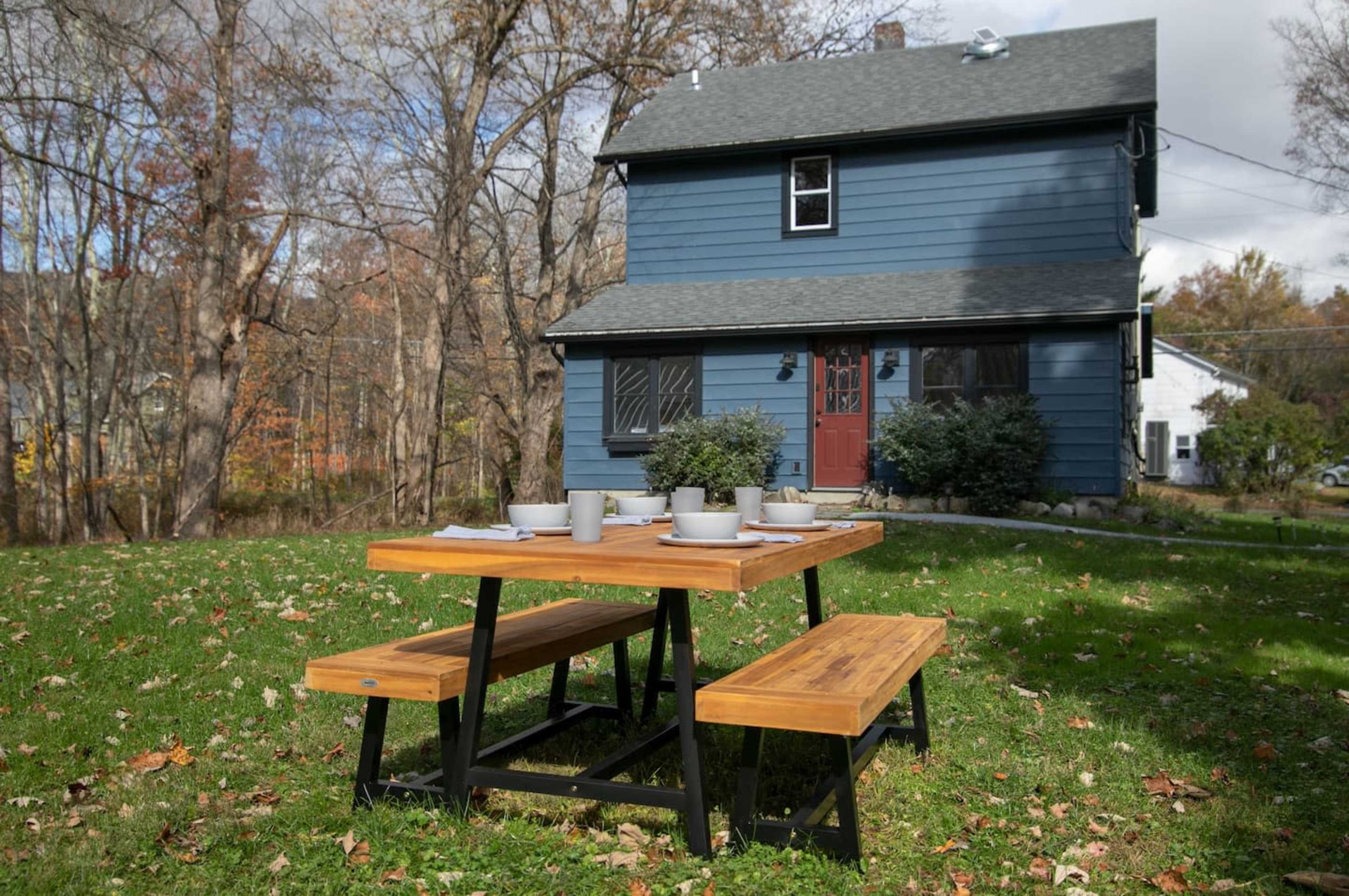 A wooden picnic table with benches is set up on the grass in front of a blue house surrounded by trees.