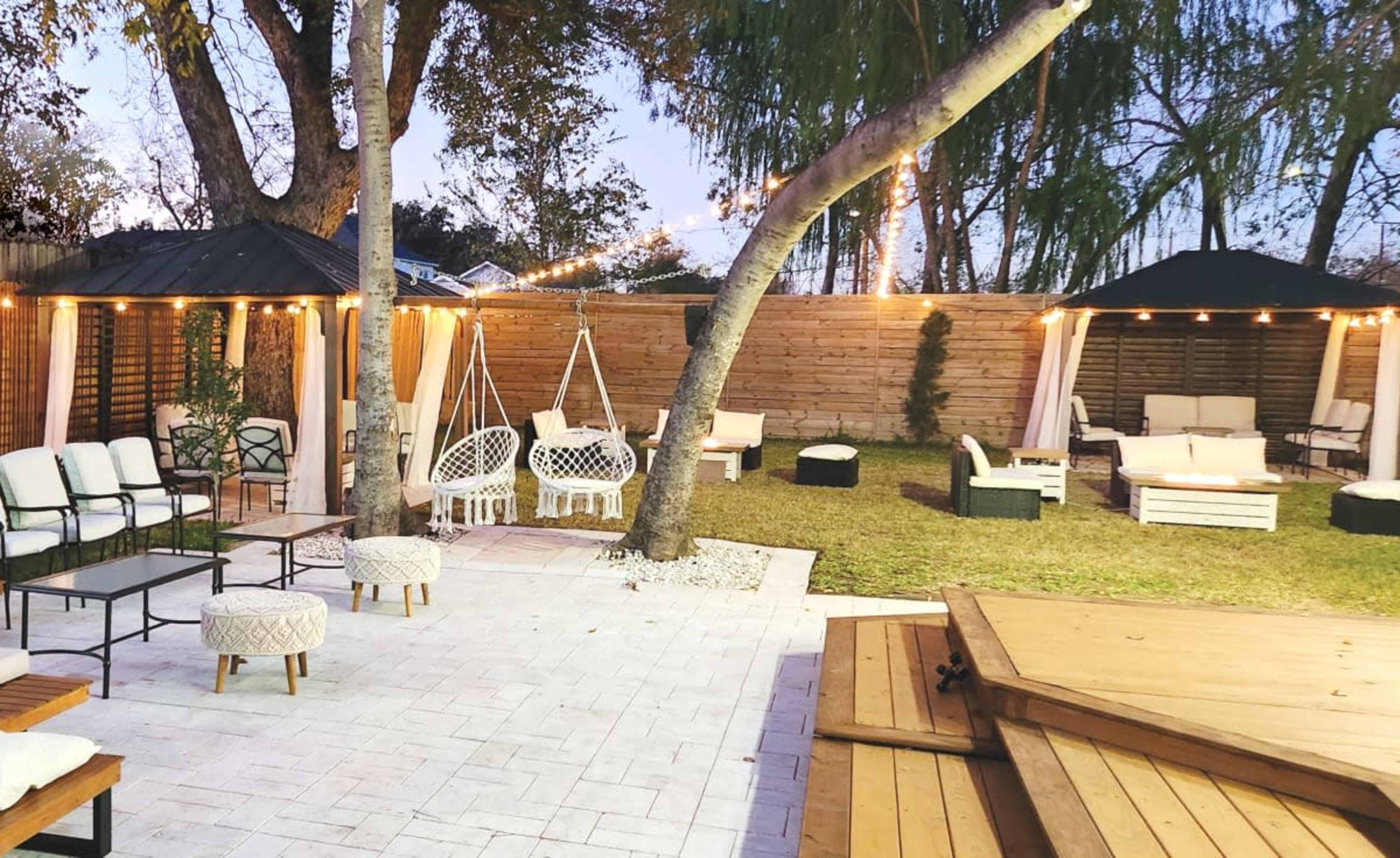 A well-decorated backyard with seating areas, hanging chairs, and canopies illuminated by string lights.