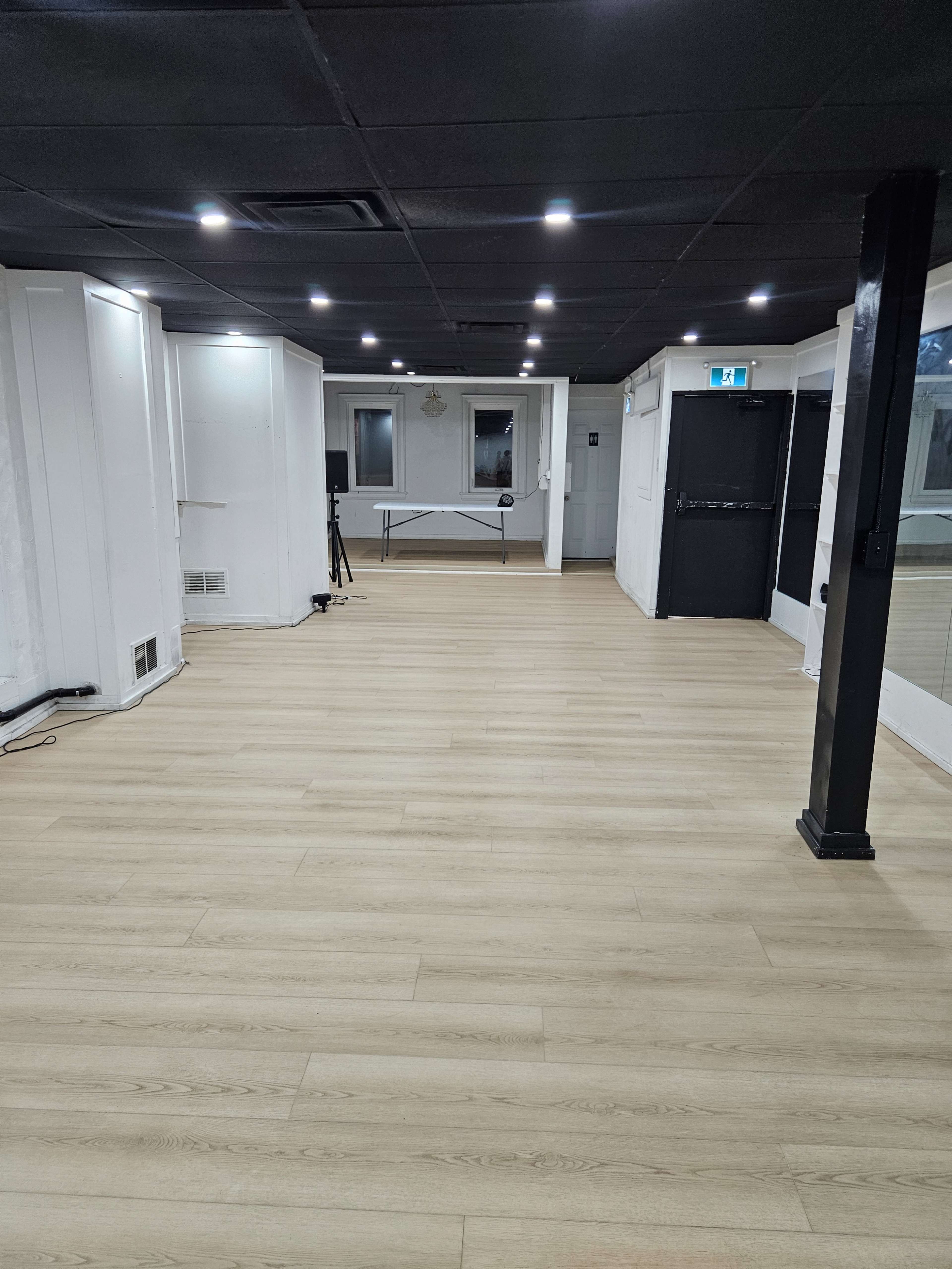 The image shows an empty room with light-colored wooden flooring, white walls, and black ceiling tiles, featuring a folding table at one end and a door leading outside.