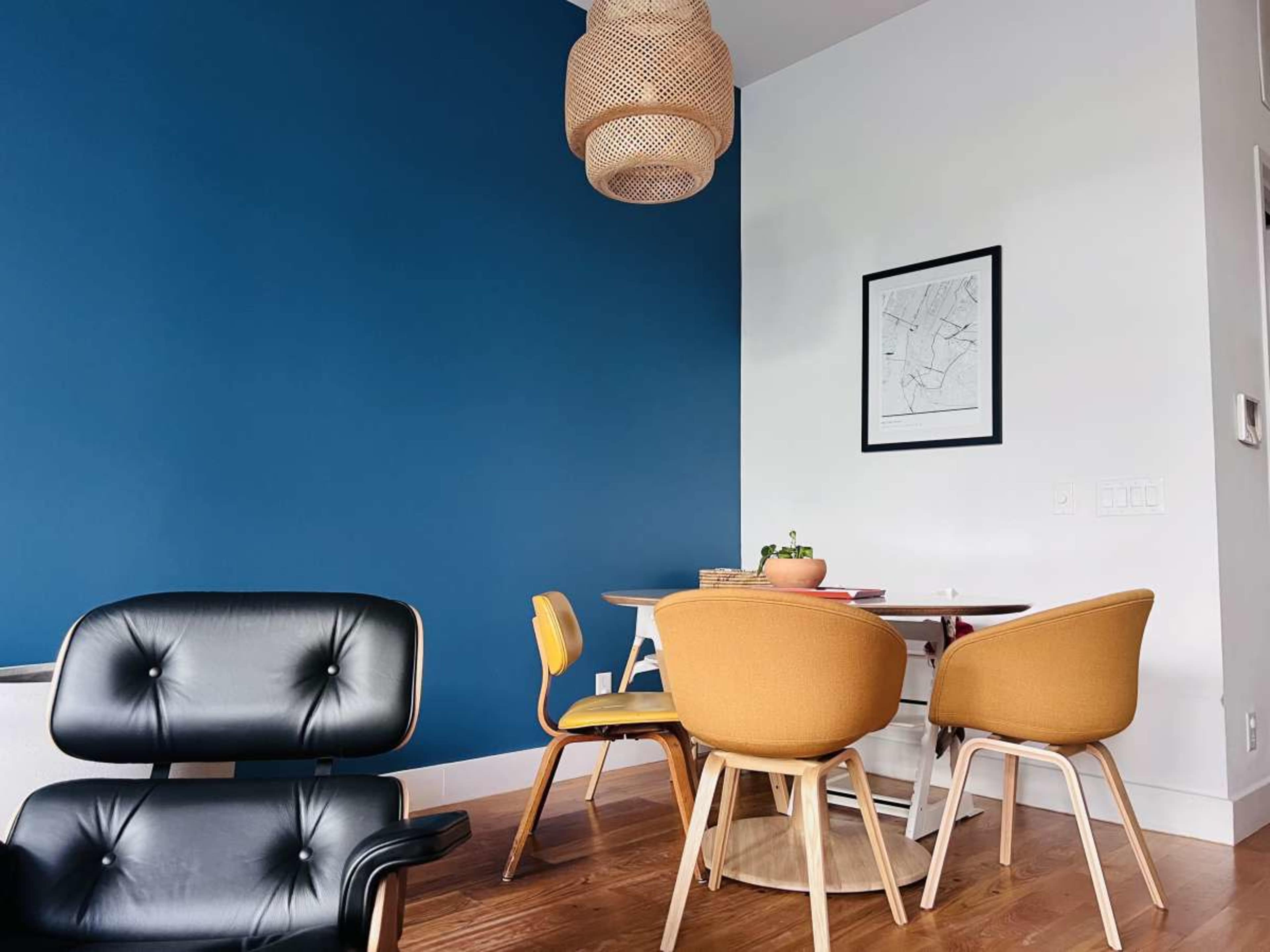 A modern dining area features a round table surrounded by mustard yellow and black chairs, with a blue accent wall and a woven light fixture overhead.