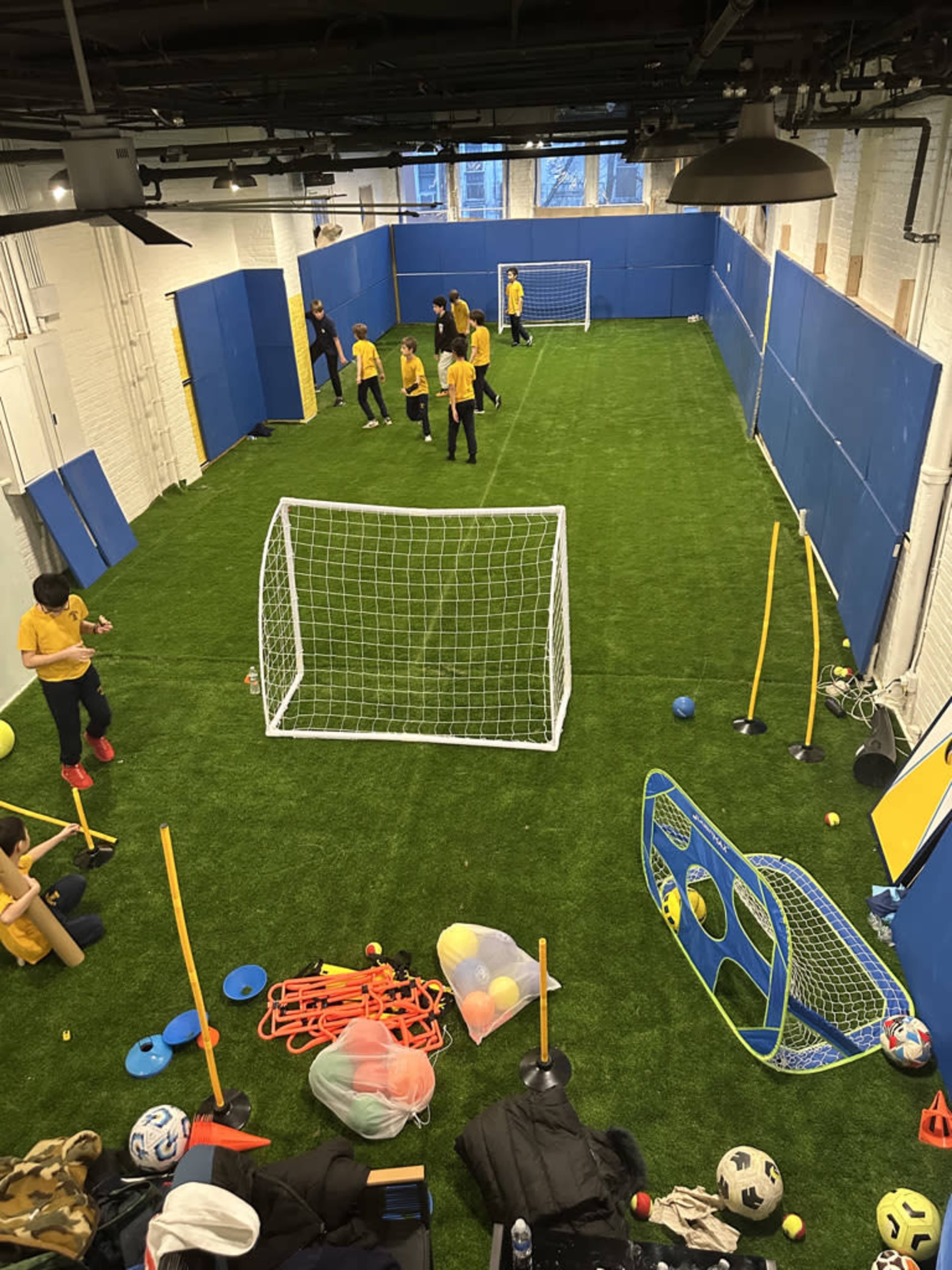 A group of individuals in yellow shirts is playing soccer on a small artificial turf field enclosed by blue walls and equipped with various sports equipment.