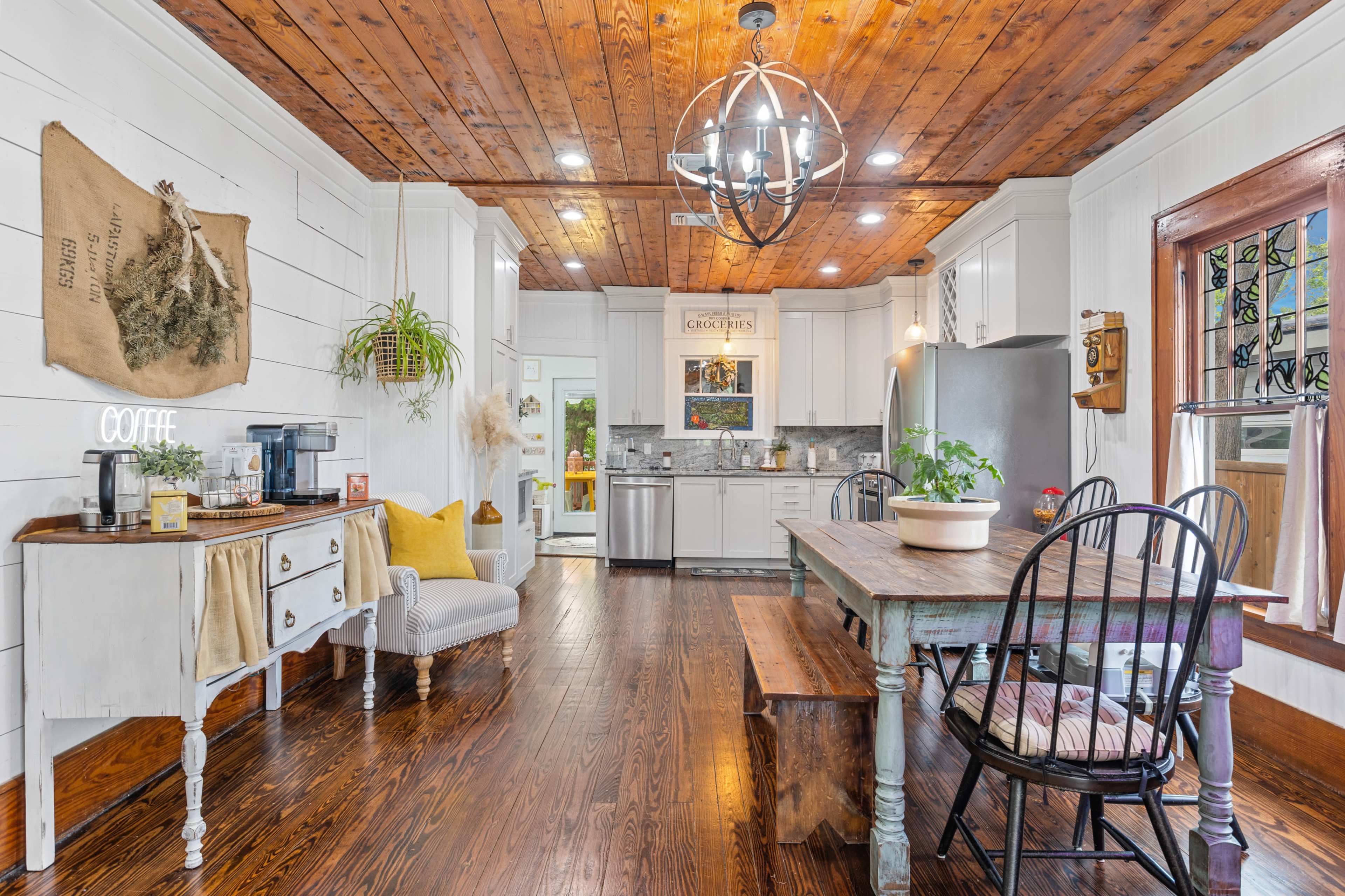 A rustic kitchen features a wooden ceiling, a dining table with black chairs, and a coffee station beside a small seating area.