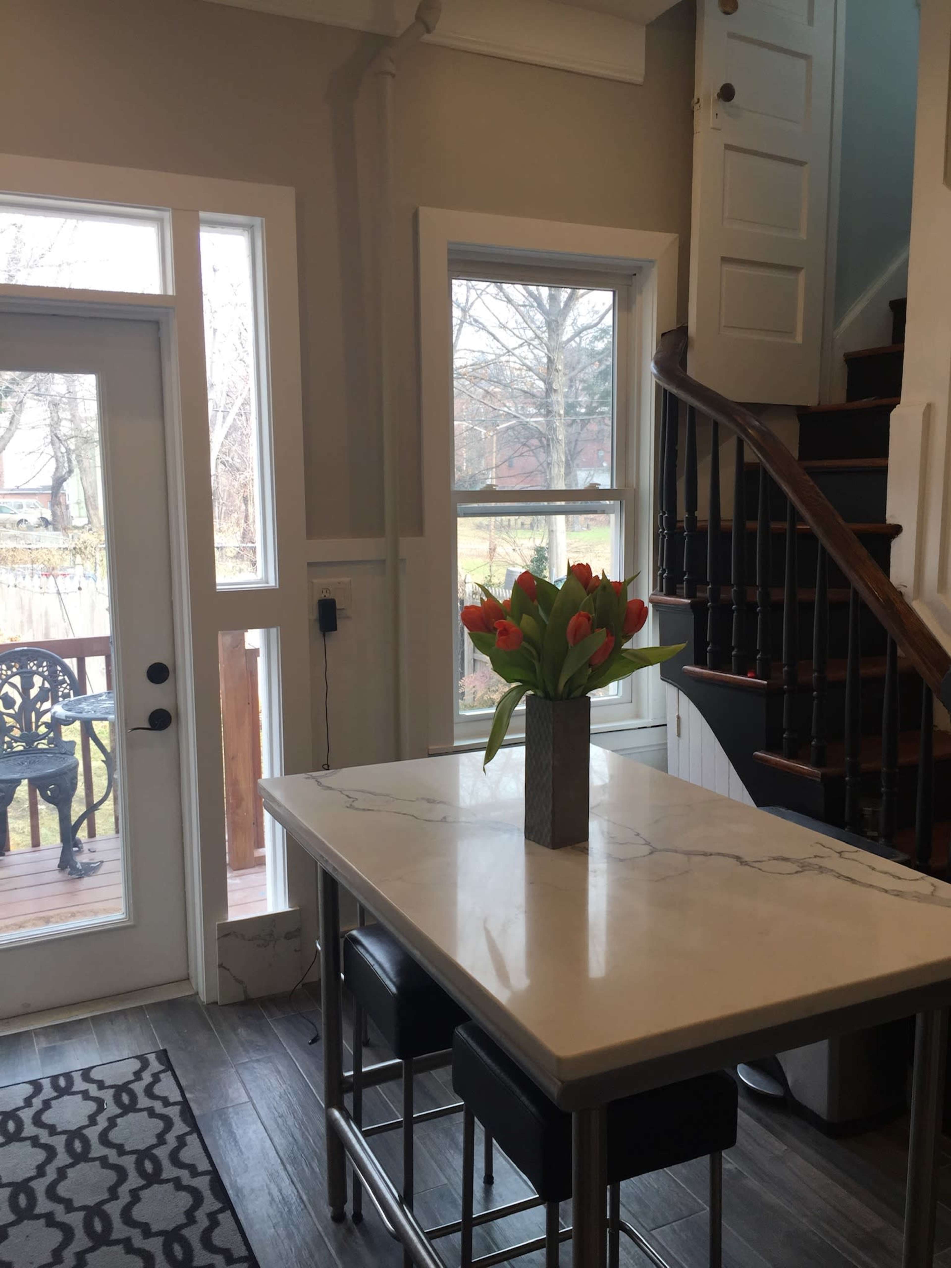 The image shows a kitchen area featuring a large table with a bouquet of red tulips, large windows, and a staircase nearby.