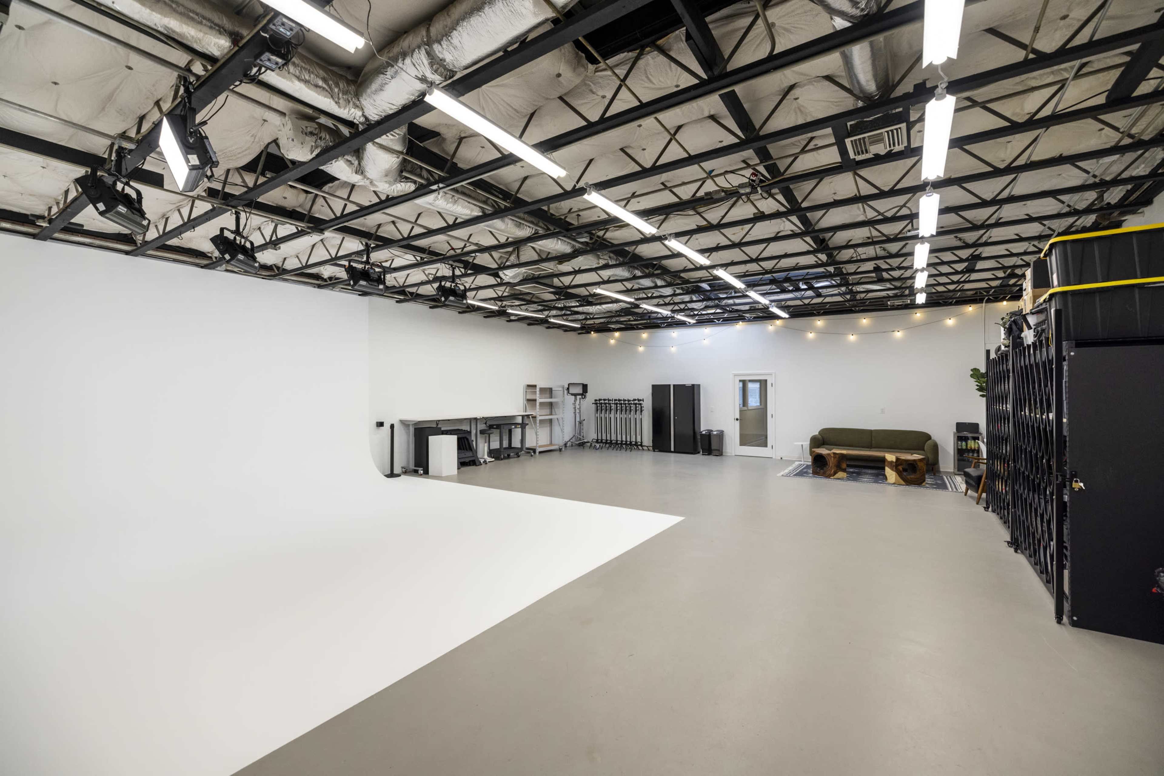 The image shows a spacious photography studio with a white backdrop, soft lighting, and various furniture and equipment arranged along the walls.