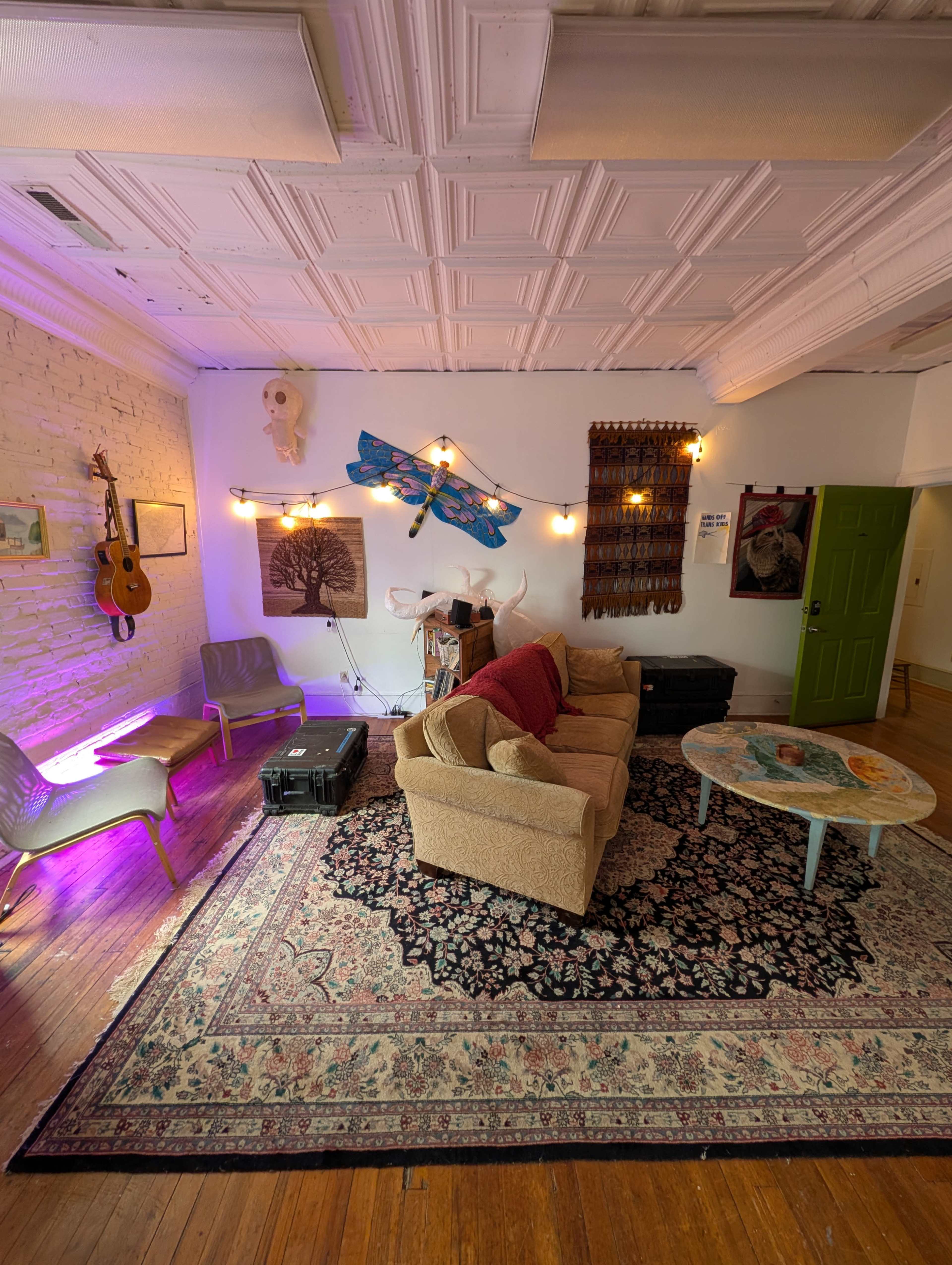 The image shows a cozy lounge area with a beige couch, two gray chairs, a round table, and various artworks displayed on the walls, illuminated by string lights.