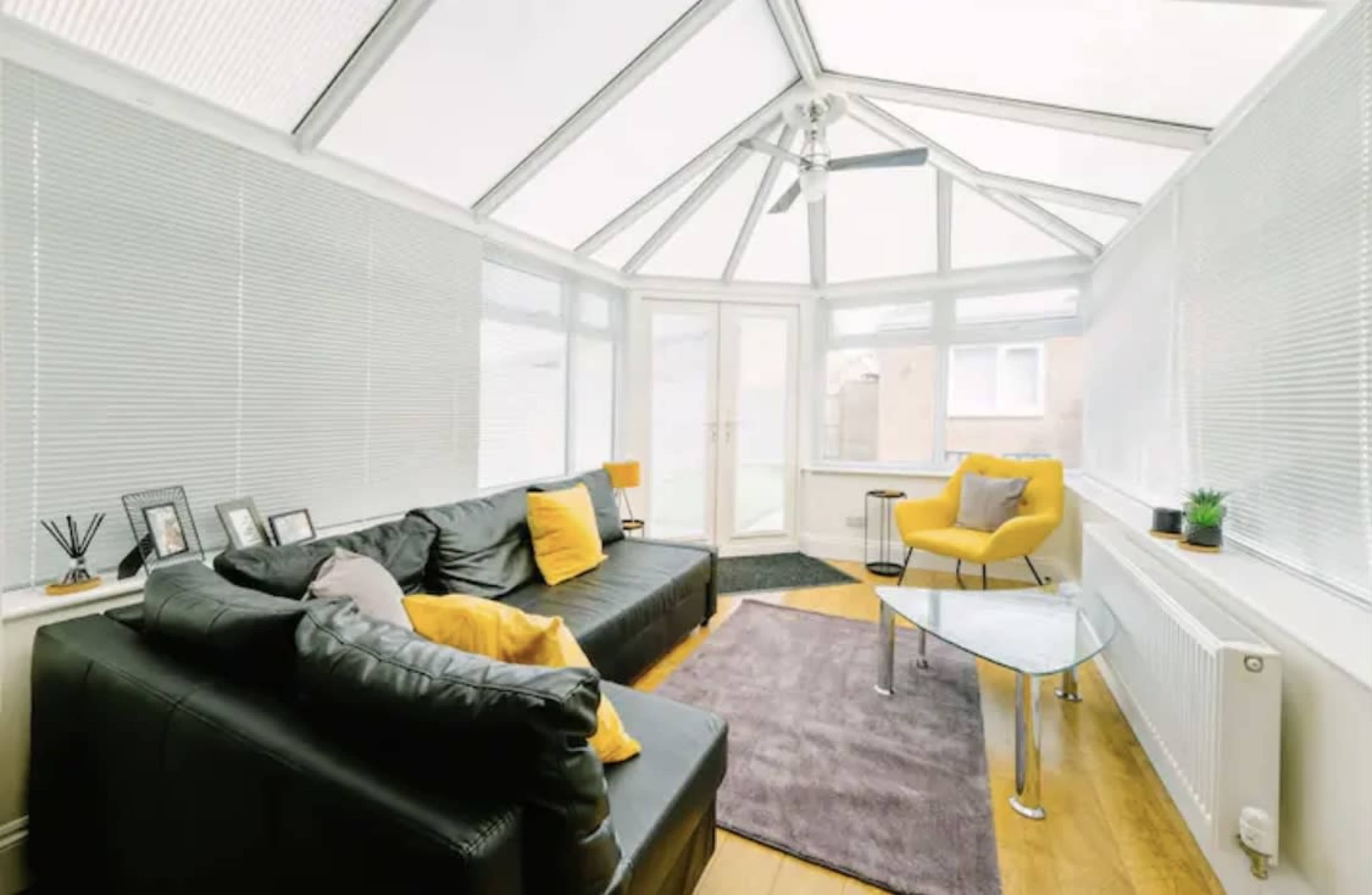 A bright conservatory with a black leather sofa, a yellow accent chair, and a glass coffee table on a rug.