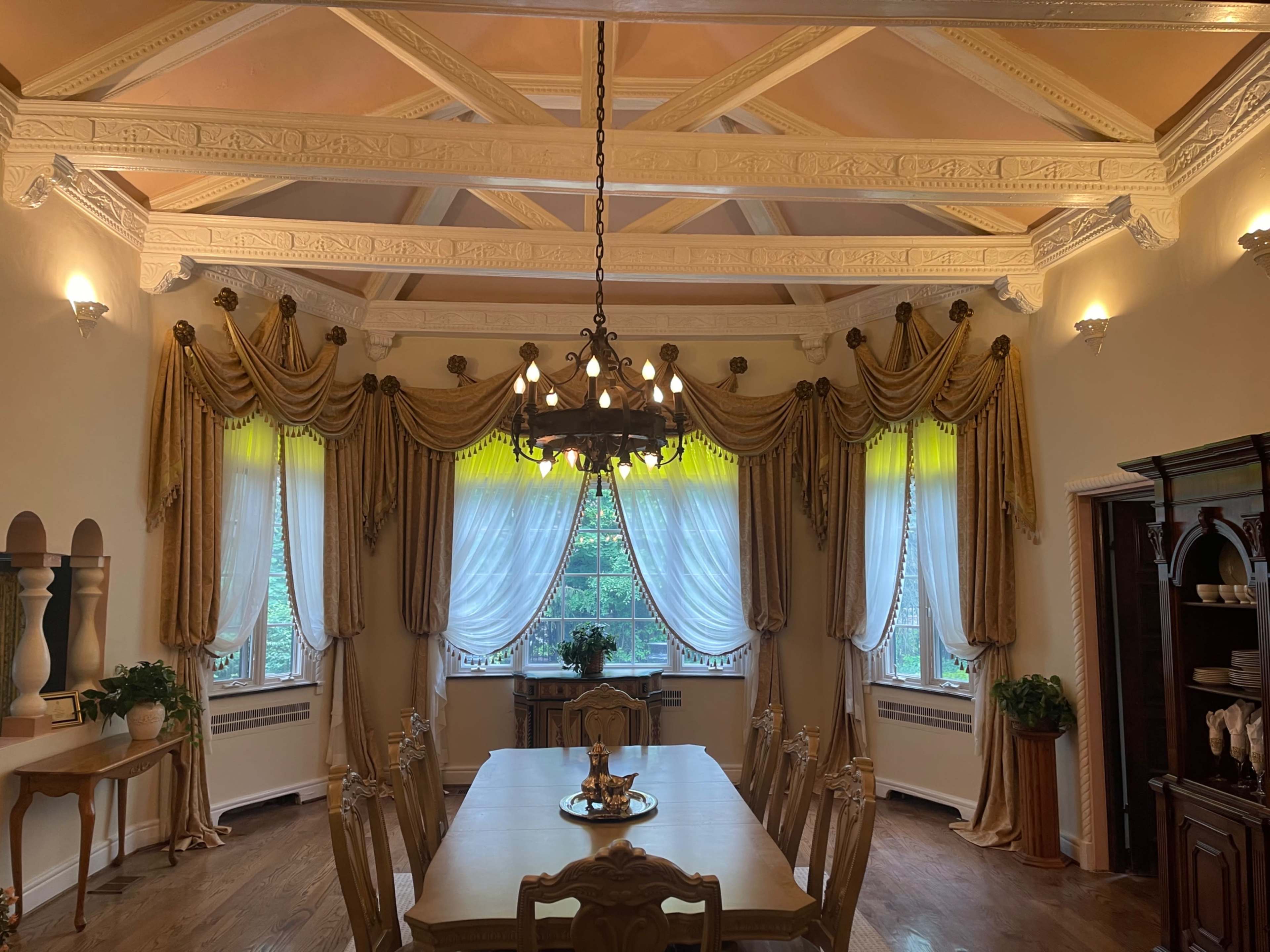 The image shows a dining room with a large table, ornate ceiling, decorative curtains, and a chandelier.