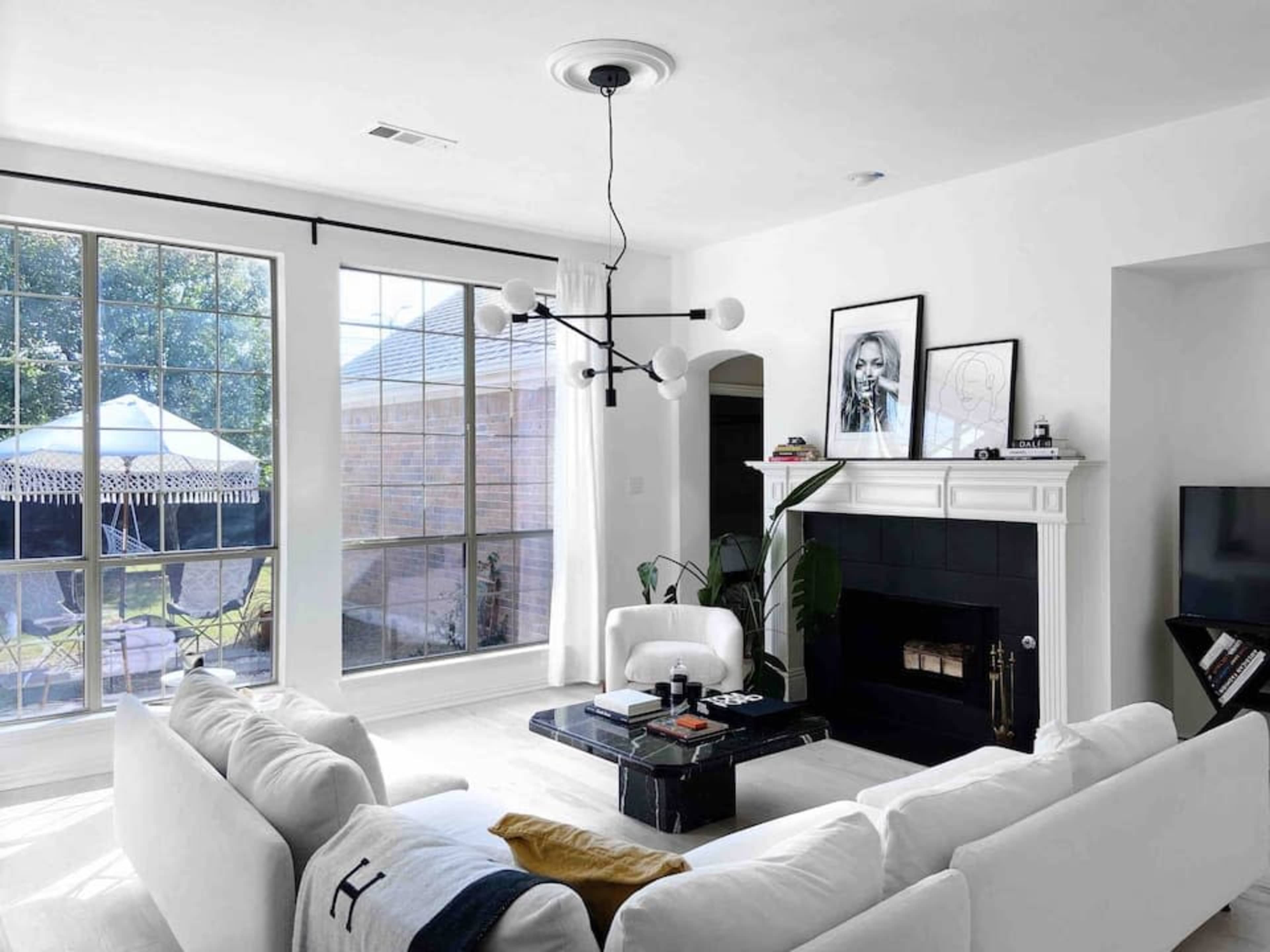 A modern living room features a white sectional sofa, a black marble coffee table, large windows with natural light, and a minimalist décor, including framed artwork and a fireplace.