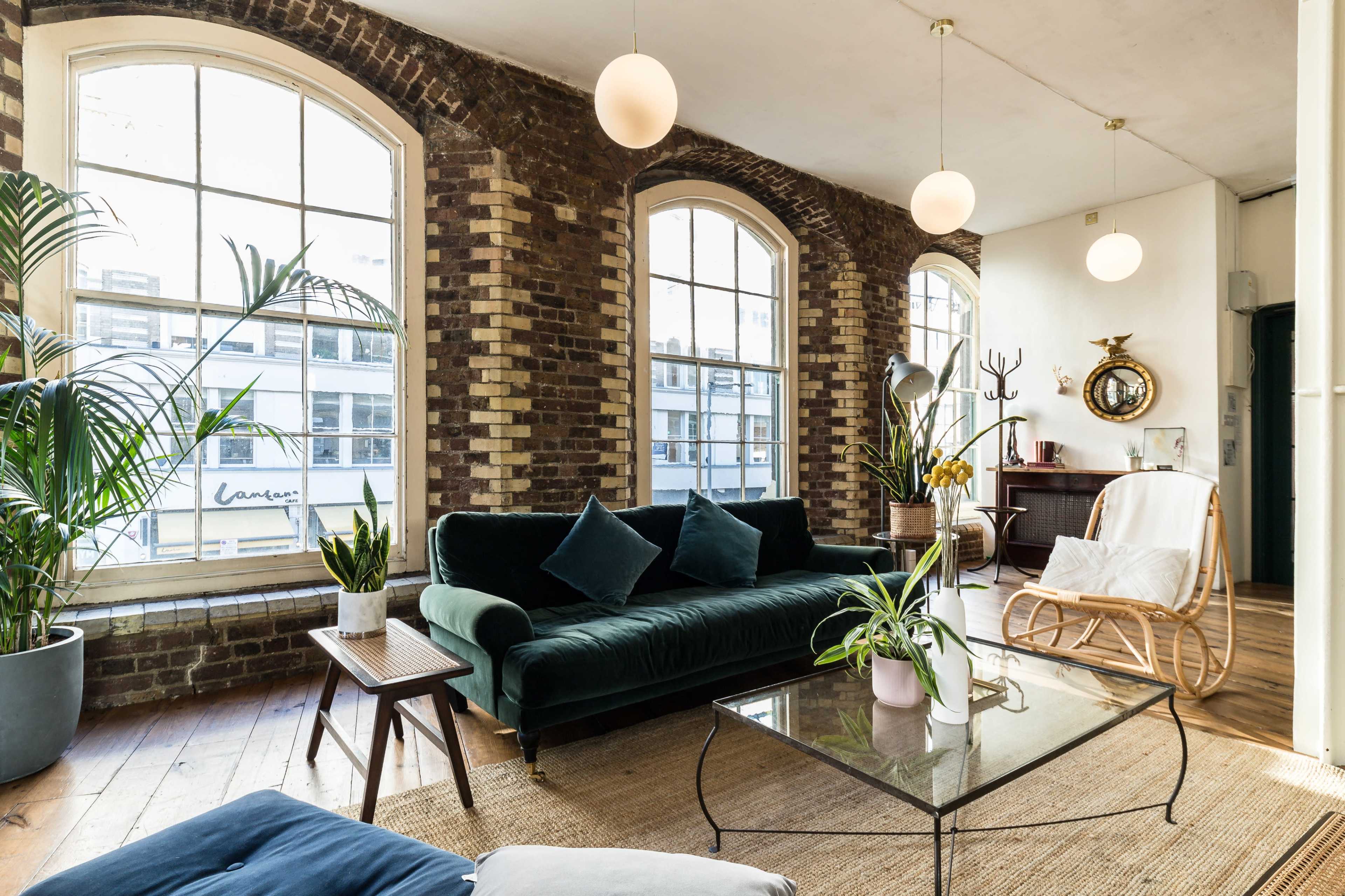 A spacious living room featuring large windows, a green velvet sofa, wooden flooring, and various plants.