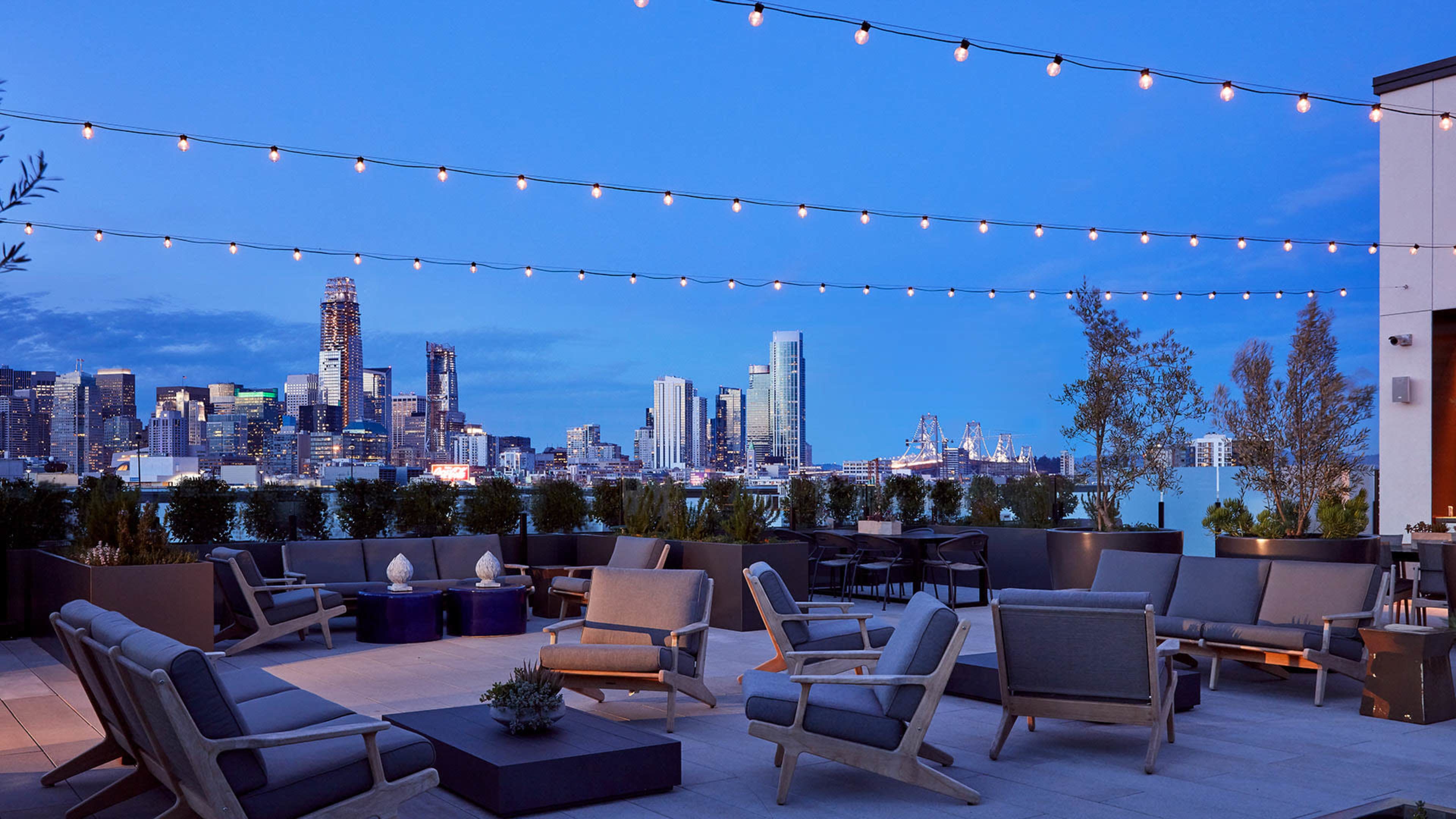 A city rooftop lounge area with string lights, modern seating, and a skyline view at dusk.