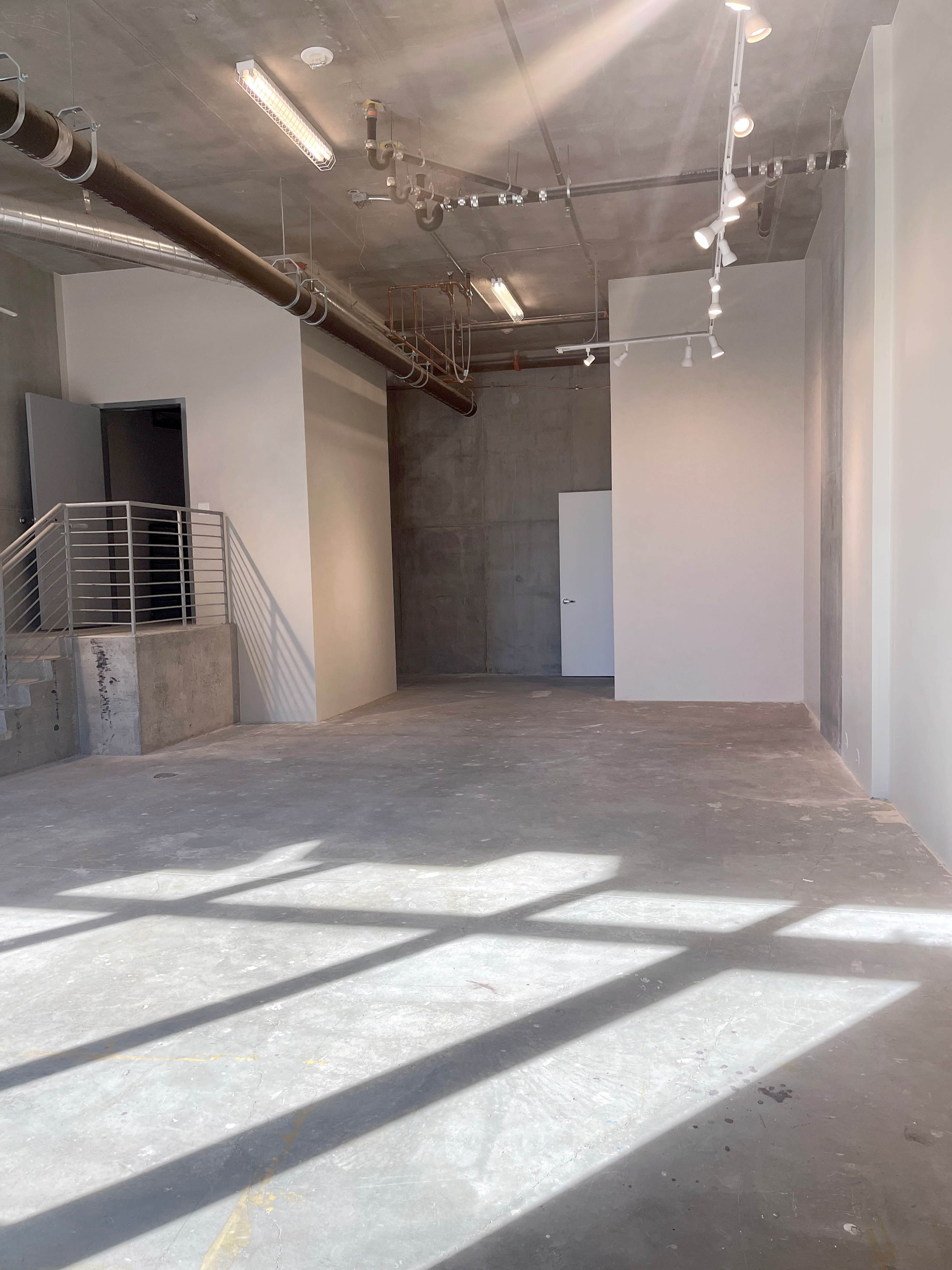 The image shows an empty, industrial-style room with concrete walls and floor, showcasing overhead lighting and a staircase leading to a doorway.