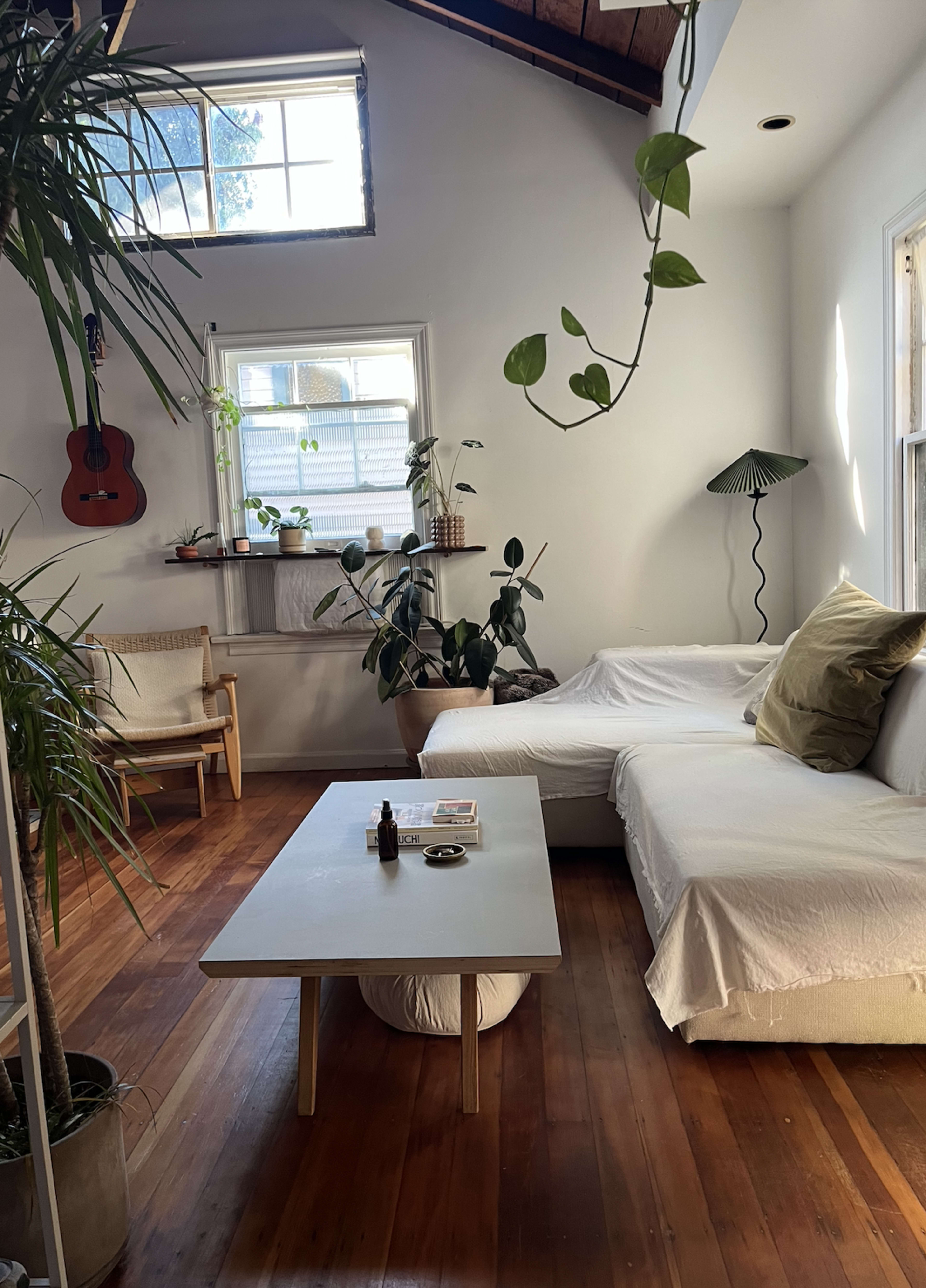A cozy room featuring a white sofa, a coffee table, various indoor plants, and a guitar hanging on the wall.