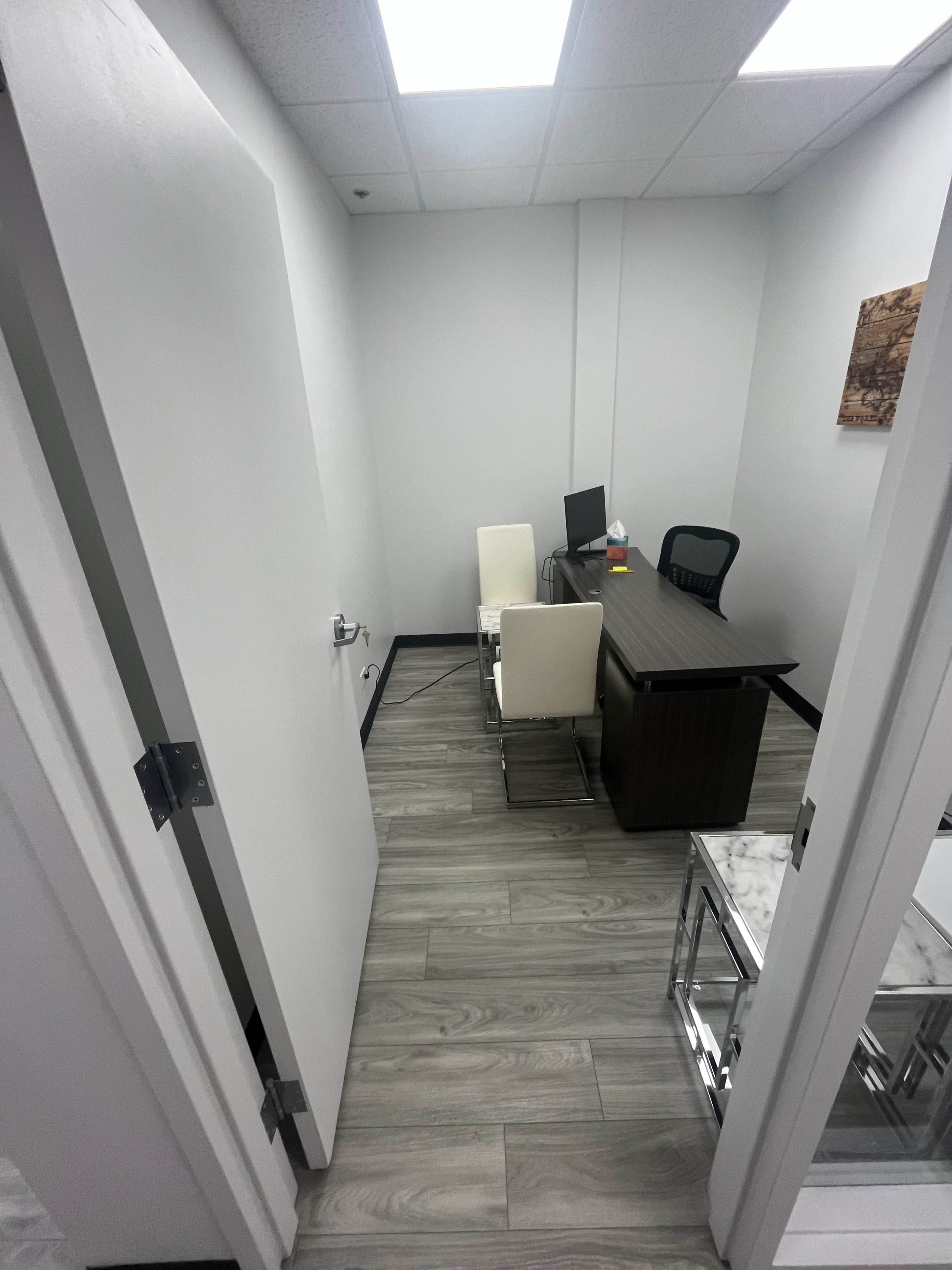 The image shows a small office with a desk, a computer, and two chairs, viewed from an open doorway.