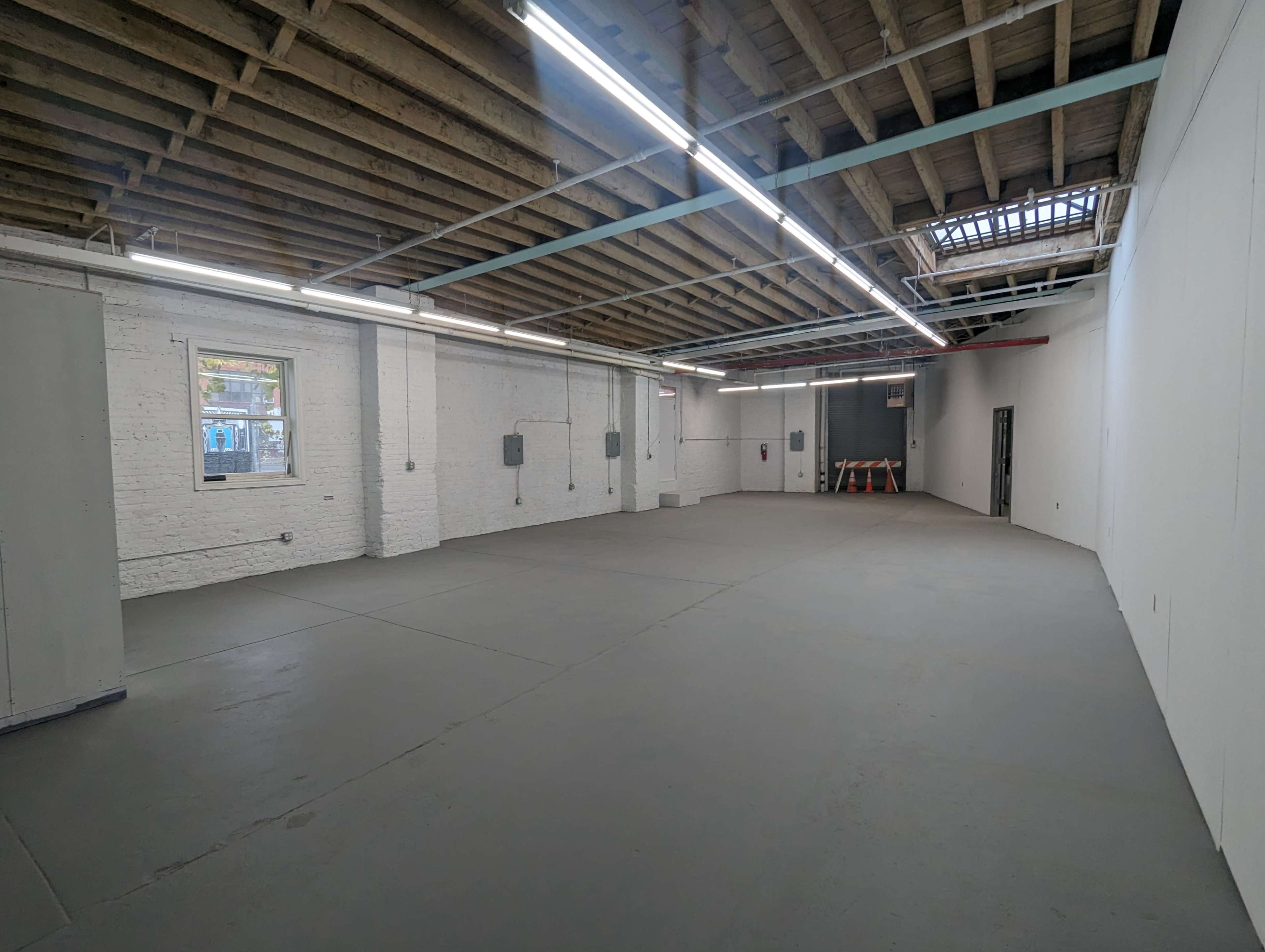 The image shows a spacious, empty warehouse with exposed wooden beams, concrete flooring, and a few electrical panels along the walls.