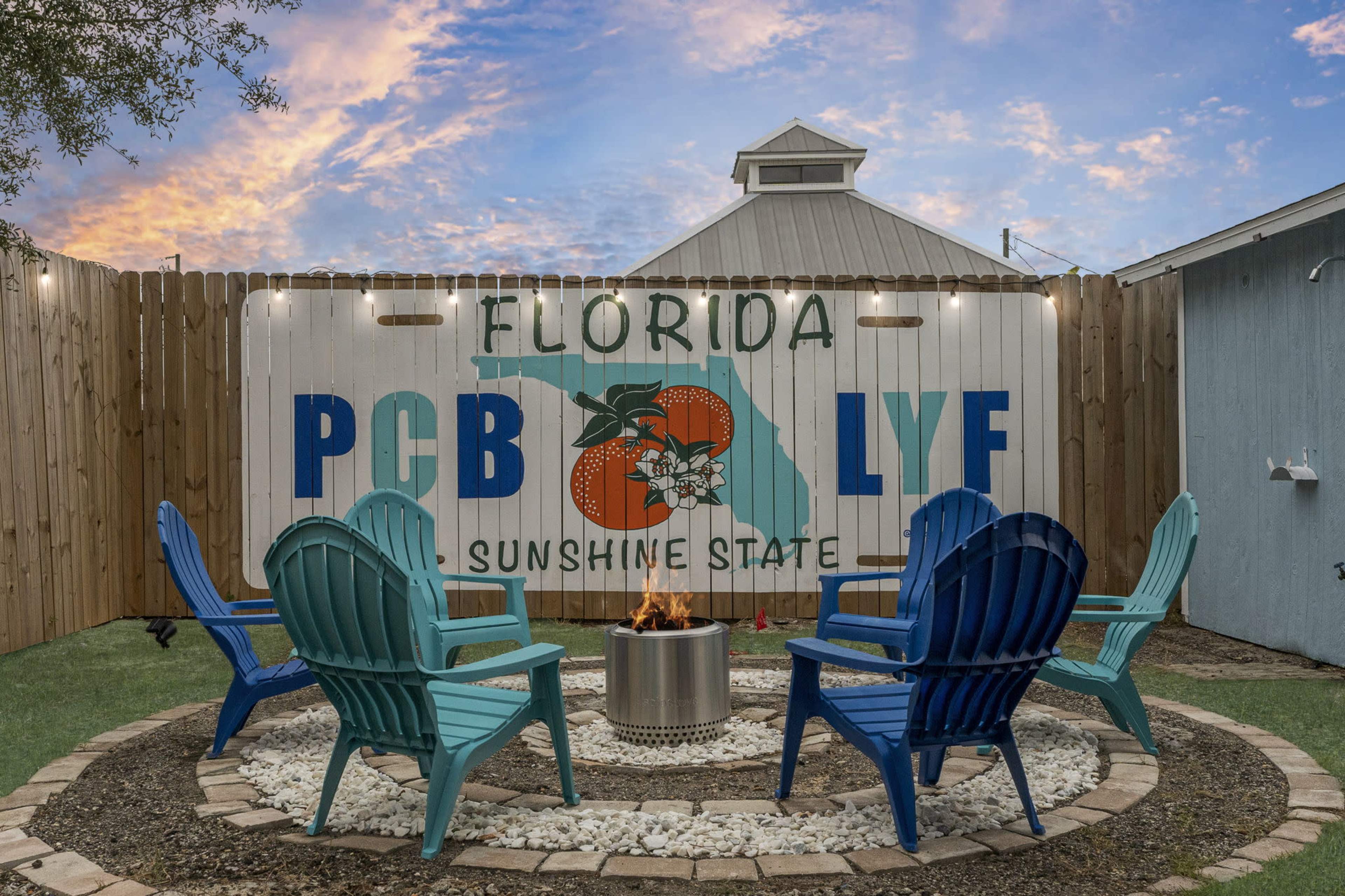 The image shows a cozy outdoor fire pit area with blue and green adirondack chairs arranged around a circular stone layout, featuring a mural of the Florida map and the words "Florida PCB LYF Sunshine State" on a wooden fence in the background.