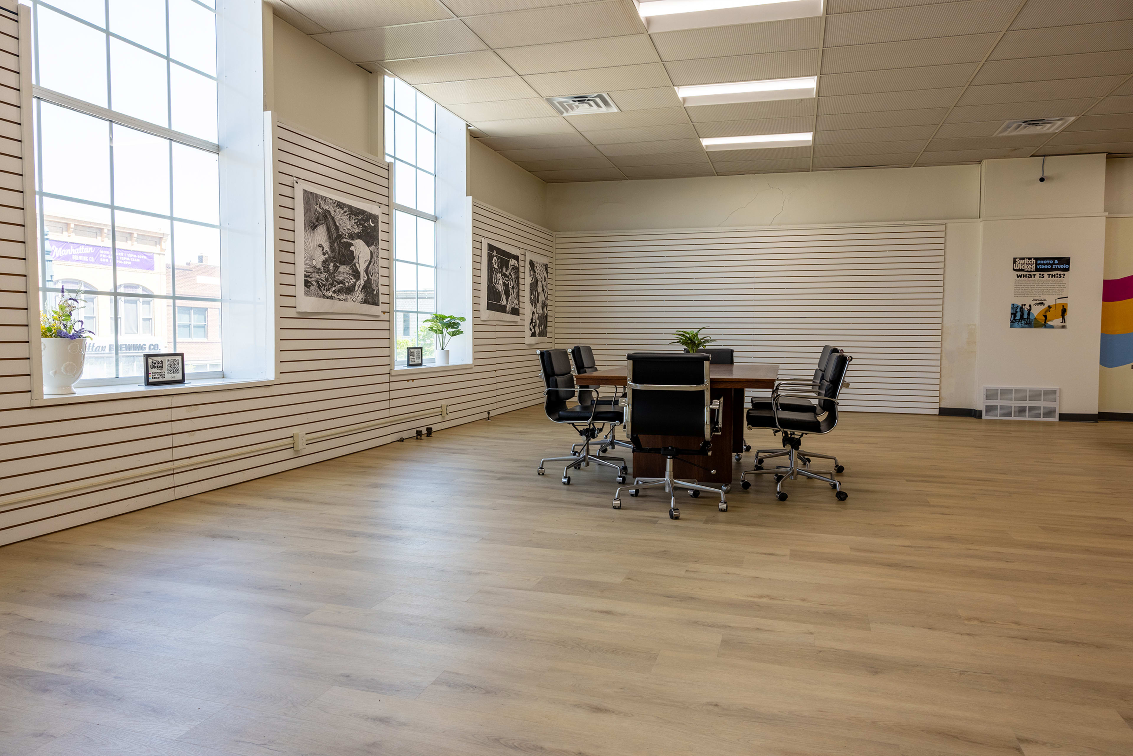 The image shows a spacious, well-lit meeting room with a large, round table surrounded by black office chairs and large windows on two walls.