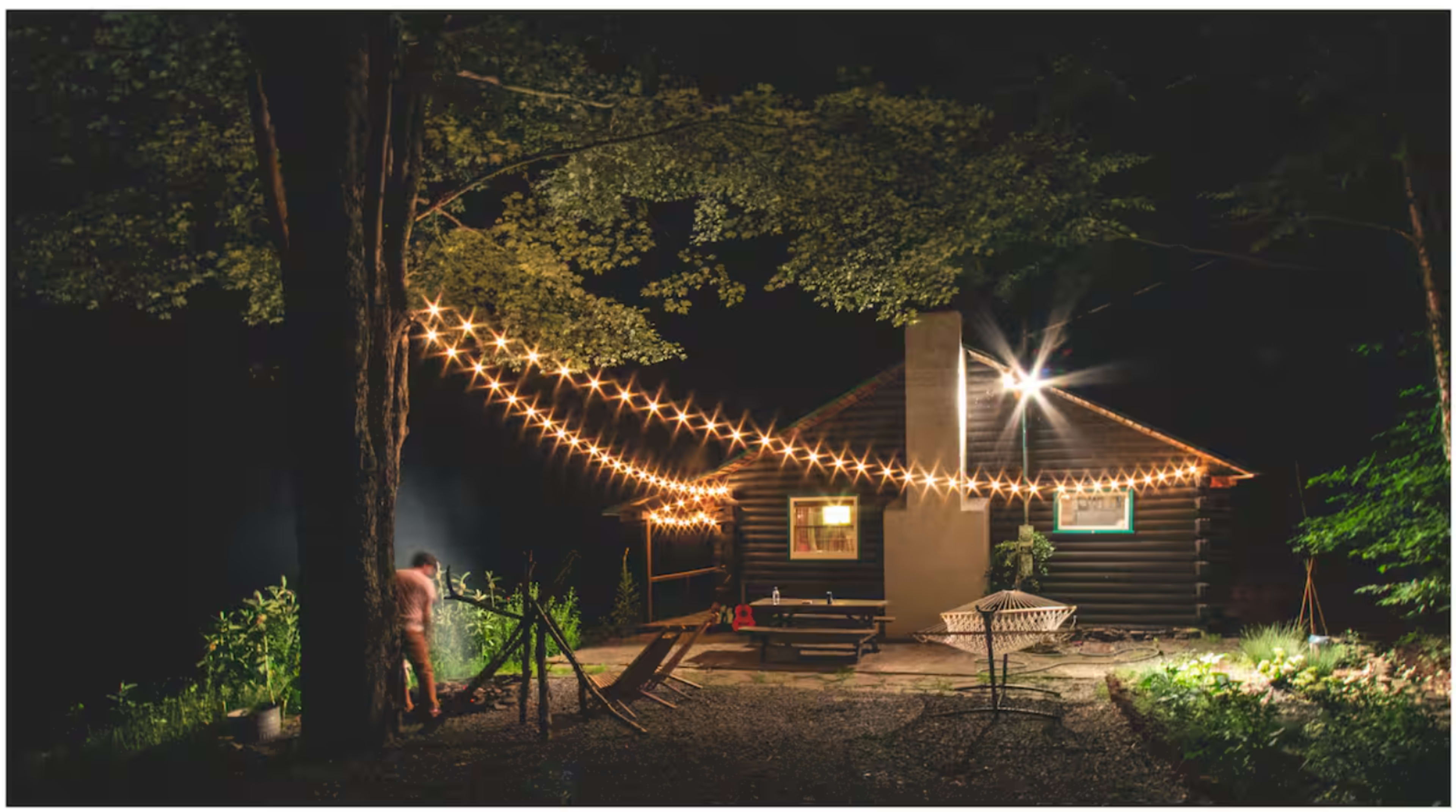 A wooden cabin is illuminated by string lights, surrounded by trees and a patio area at night.