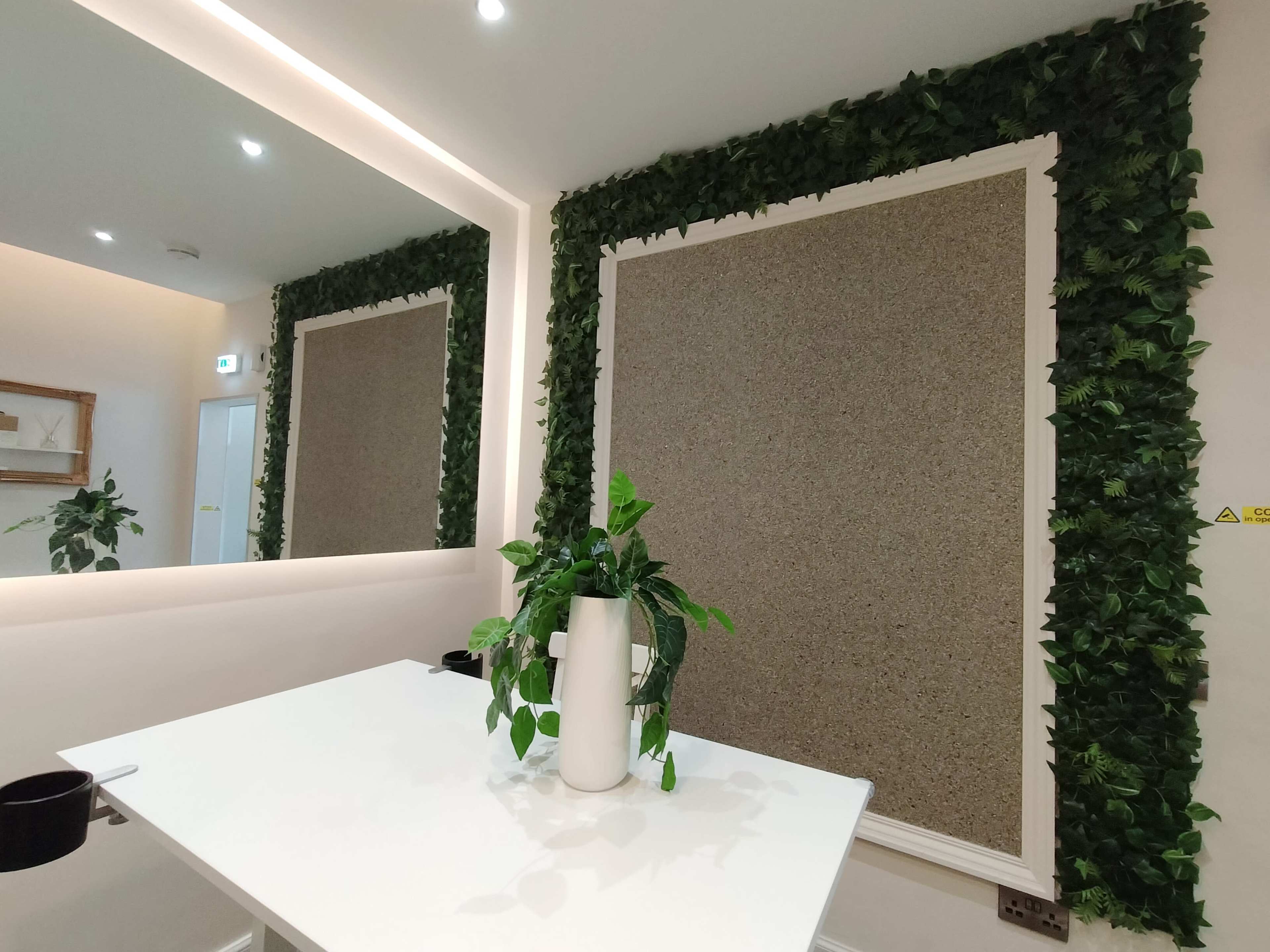 A modern interior with two cork board areas framed by green foliage, adjacent to a plain white table and a potted plant.