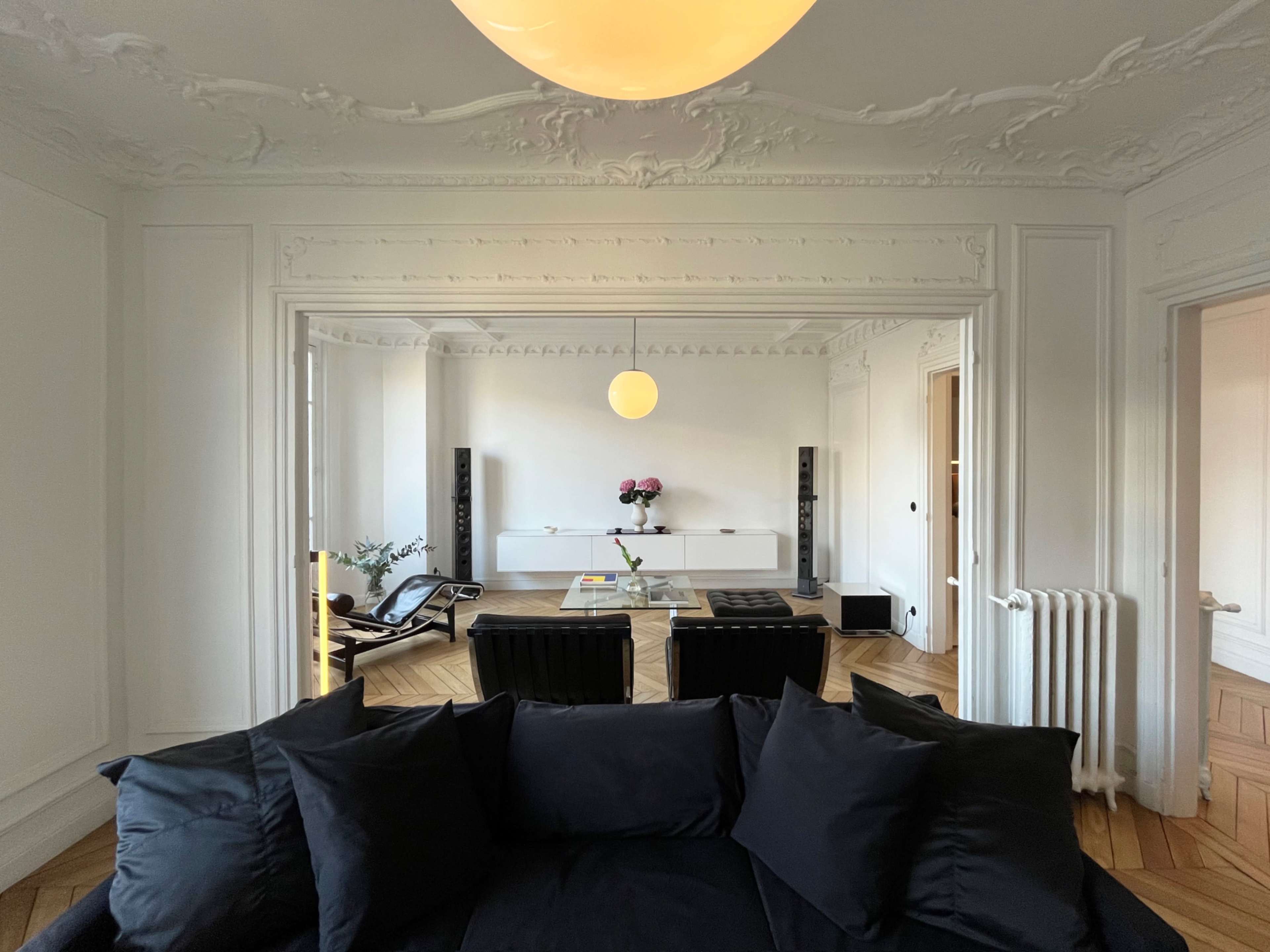 The image shows a minimalist living room with a black sofa, wooden flooring, a large central table, and decorative wall moldings.