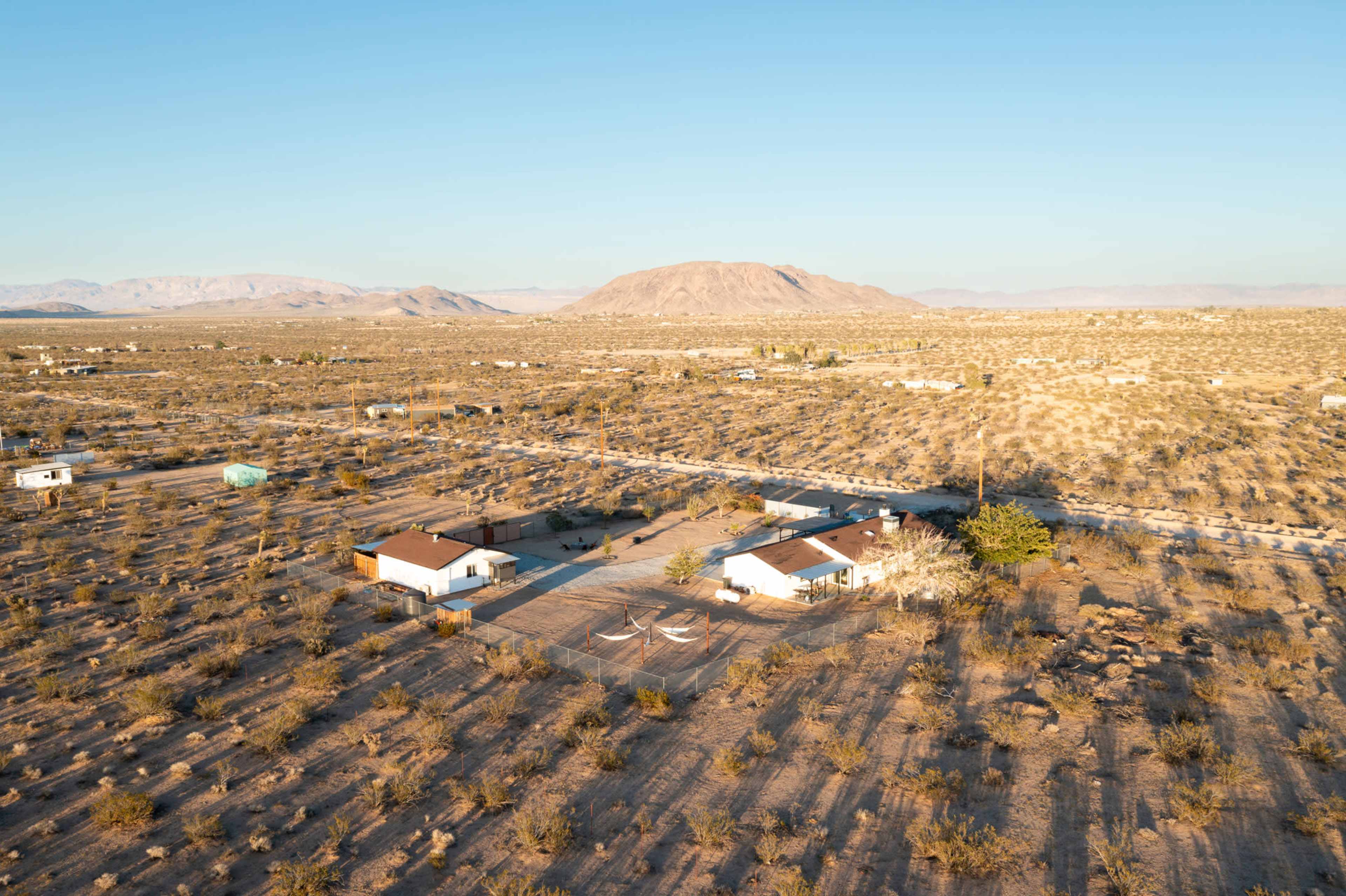 A sprawling desert landscape with a cluster of buildings, including a house and several outbuildings, set against a backdrop of distant mountains.