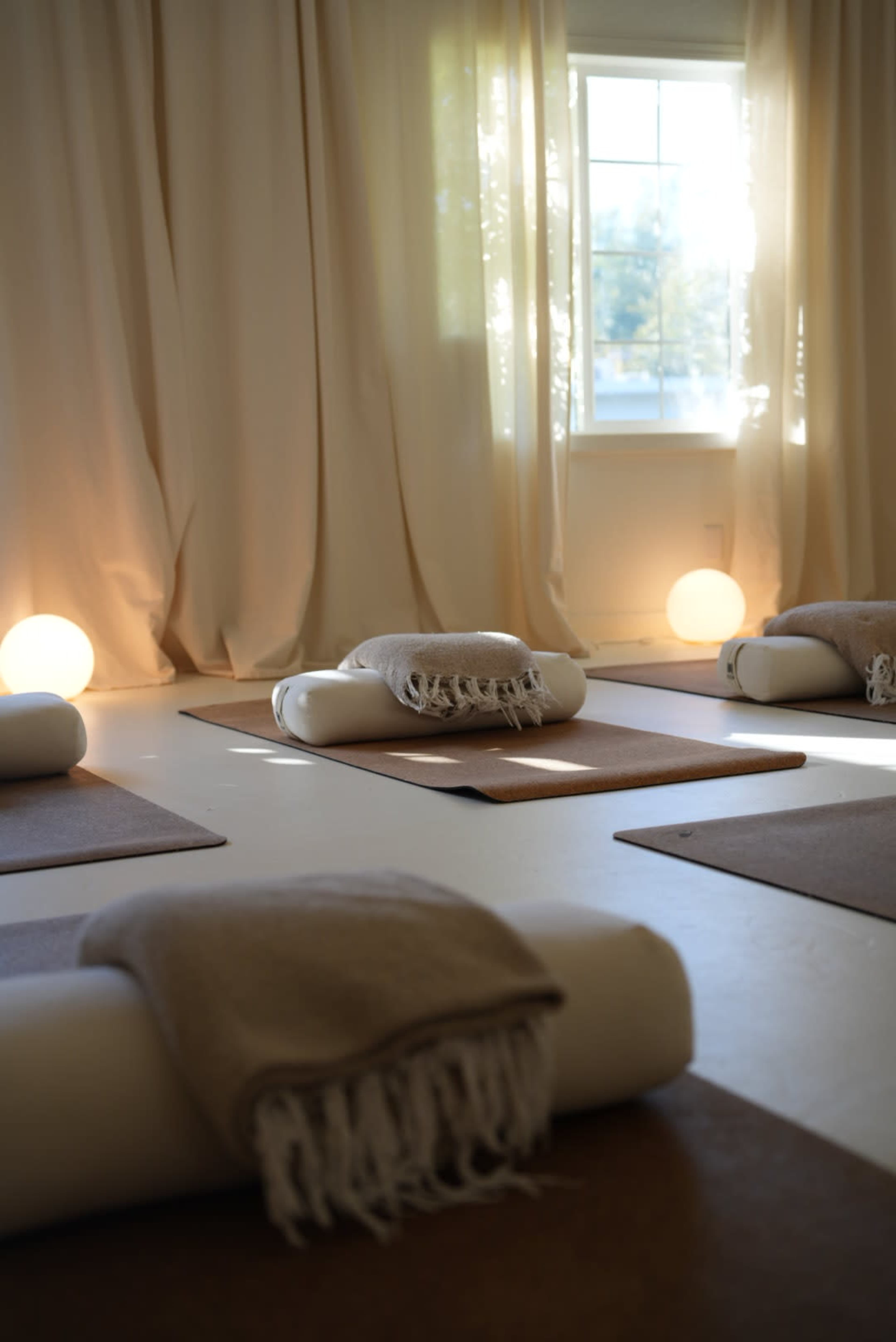 The image shows a serene yoga studio with mats and cushions arranged on the floor, illuminated by soft light from a nearby window and glowing lamps.