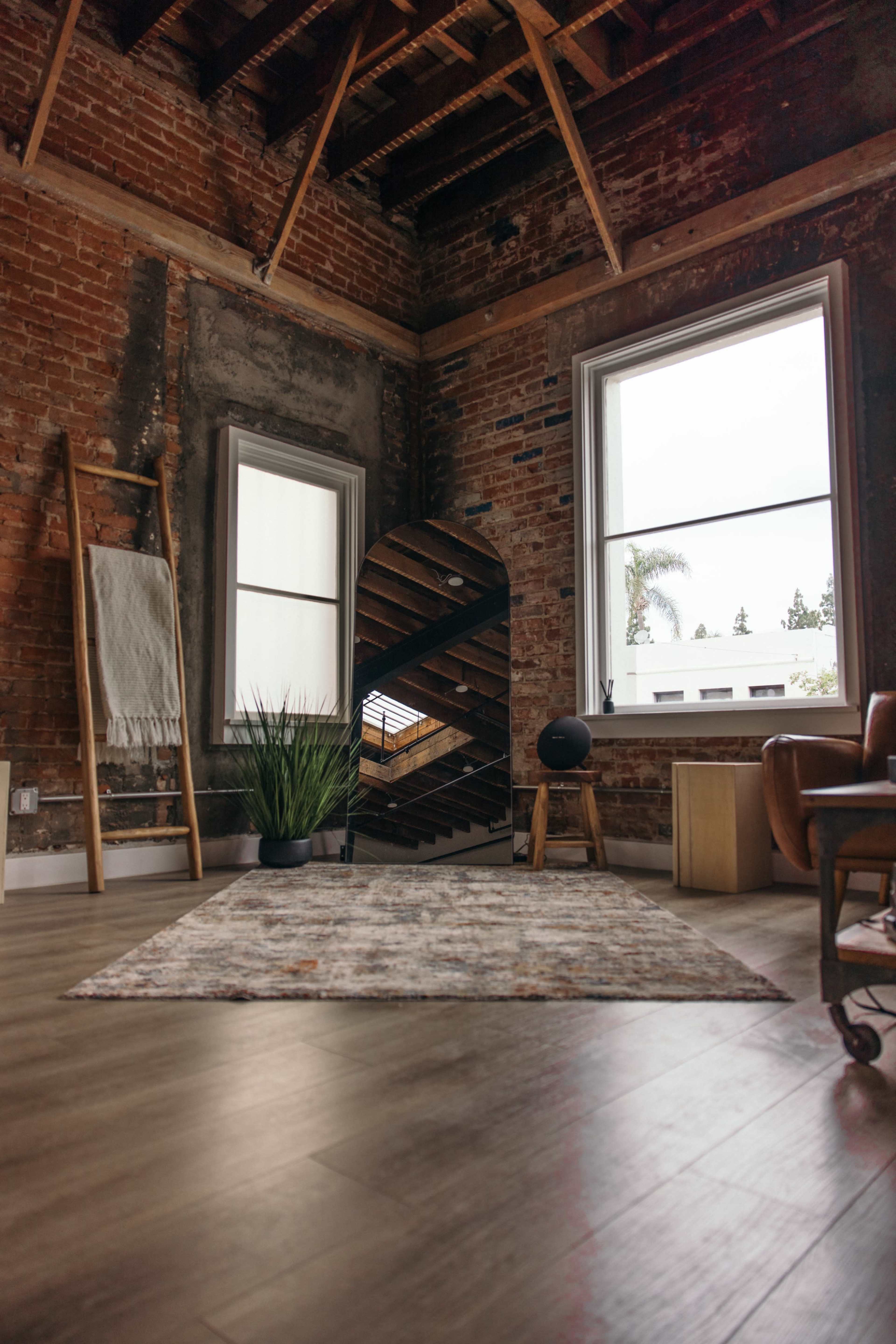 A corner of a rustic room features exposed brick walls, large windows, a spiral mirror, a small black speaker, a simple wooden stool, and a patterned rug.
