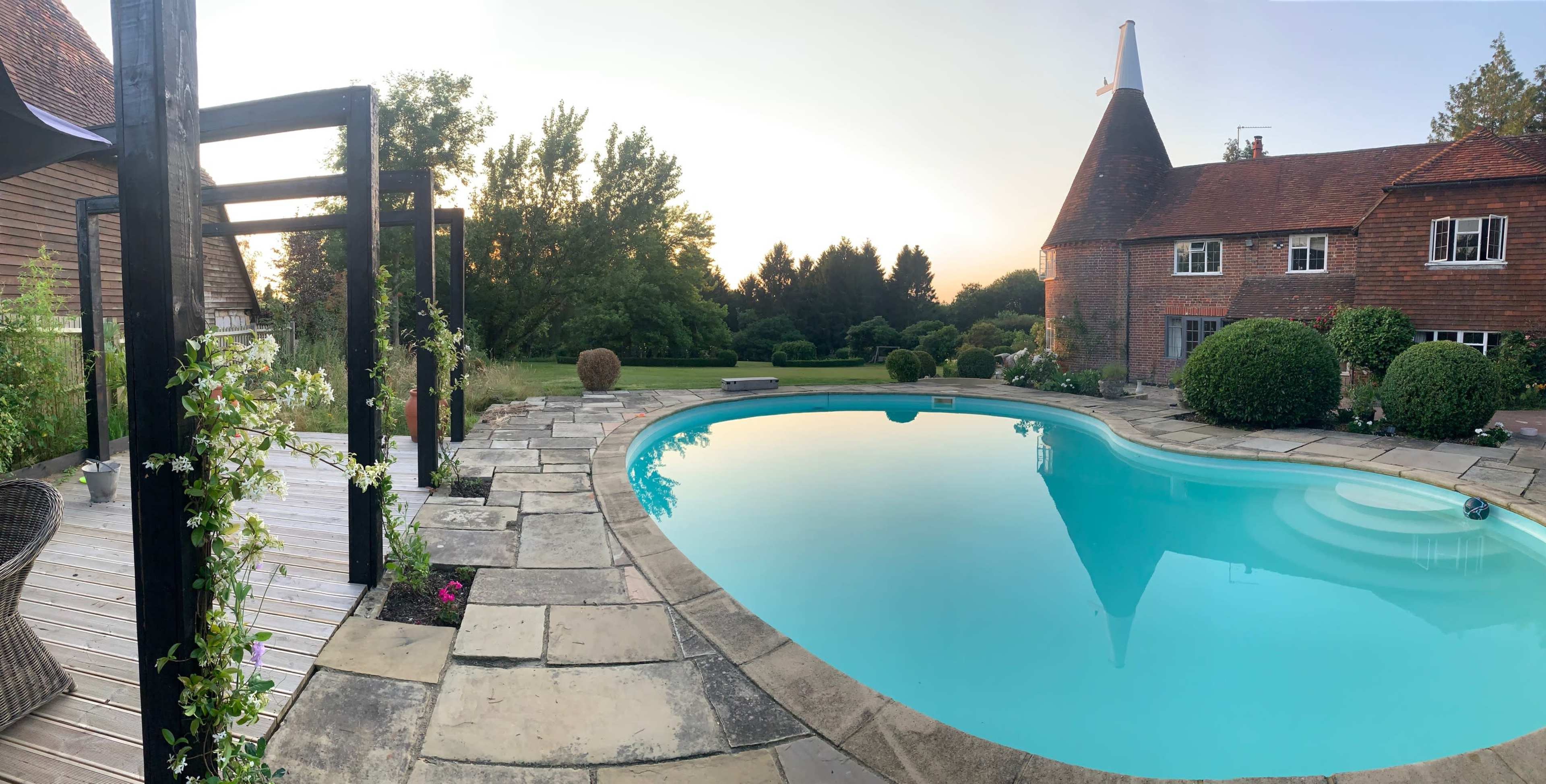 A rectangular swimming pool is surrounded by stone tiles and manicured lawns, with a wooden pergola to one side and a house featuring a unique roof design in the background.
