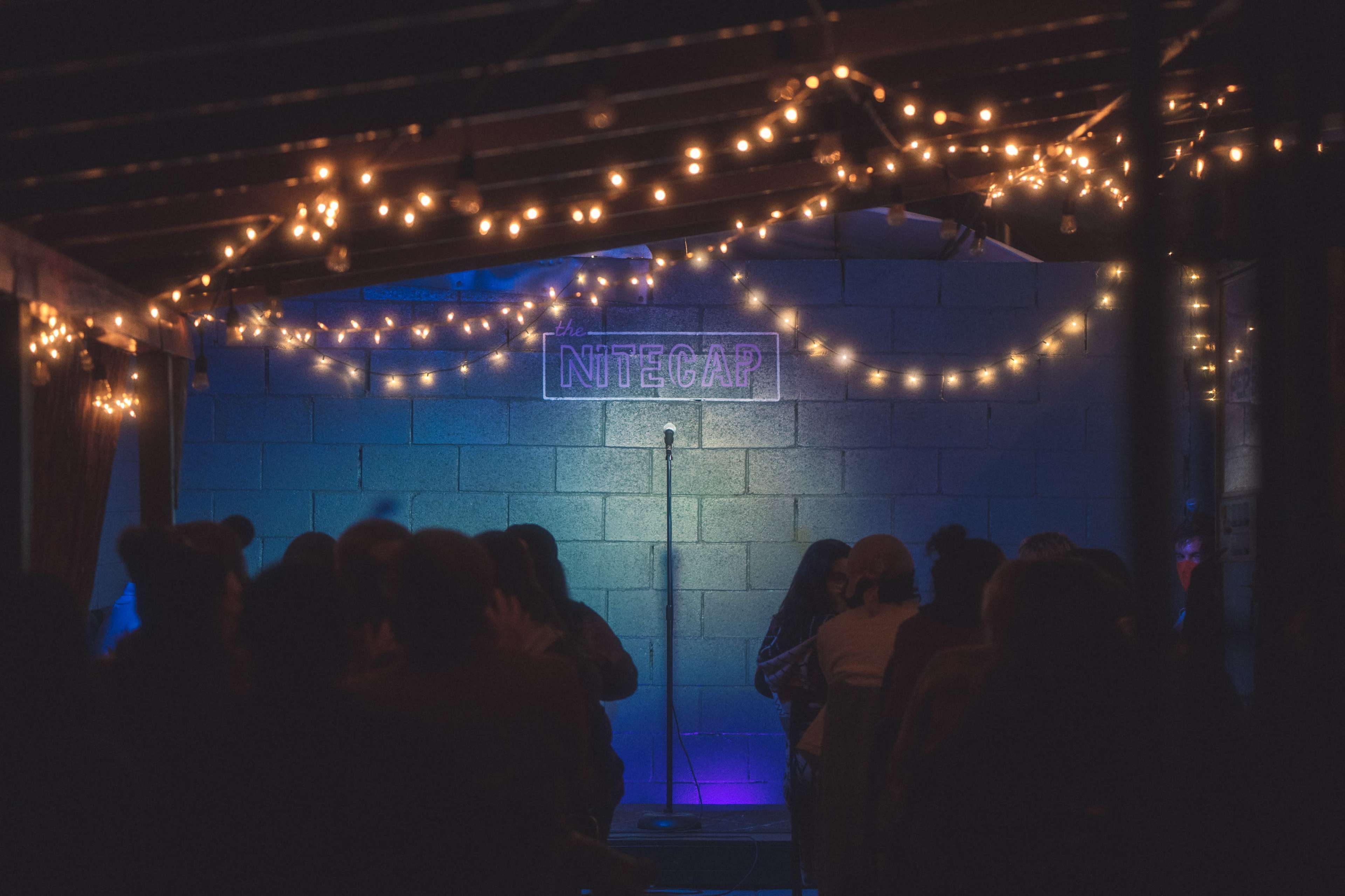 A stage with a microphone is illuminated under string lights in a dimly lit venue.