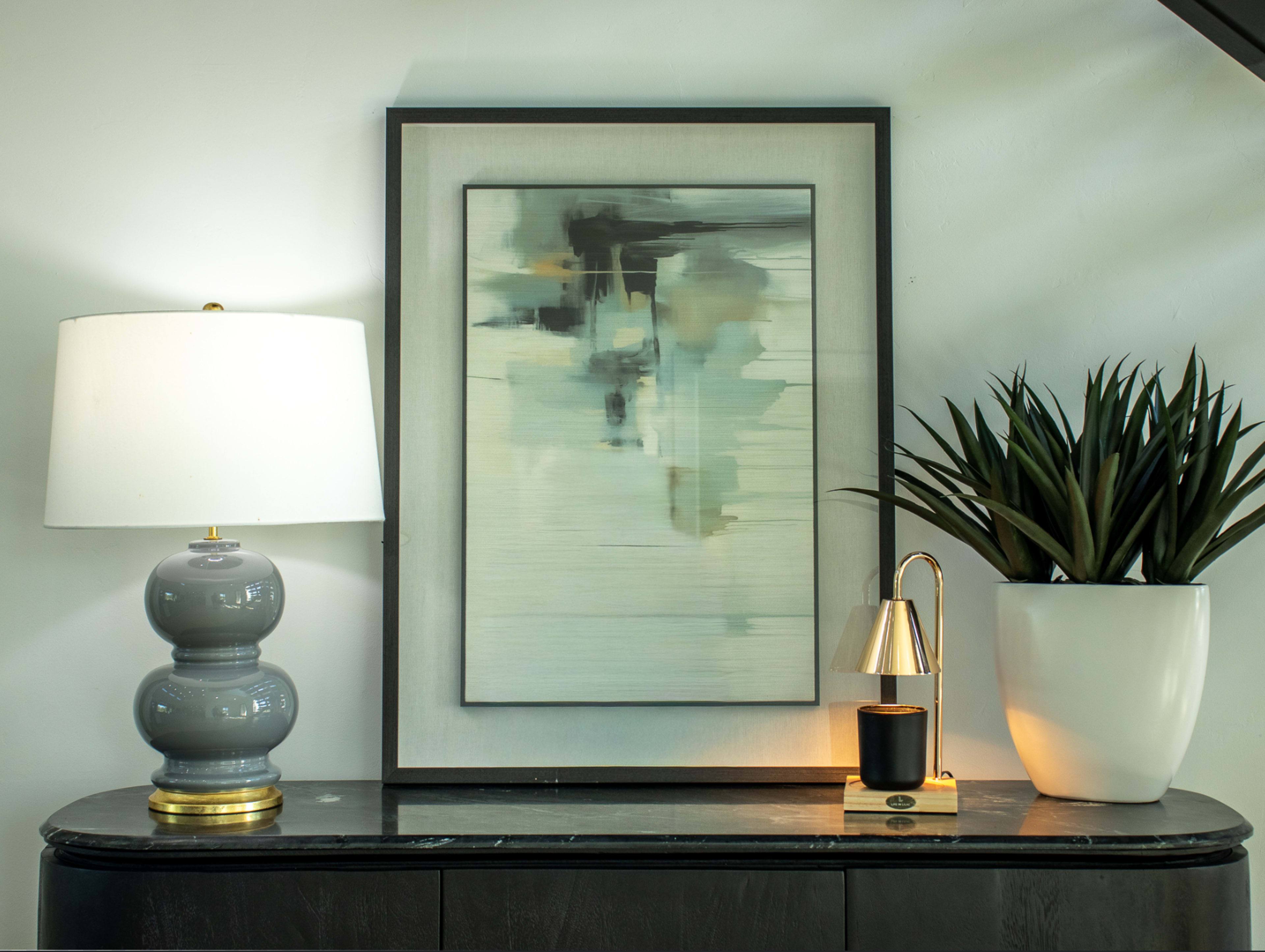 A gray lamp and a small brass lamp stand beside a large plant and a framed abstract painting on a dark wooden dresser.