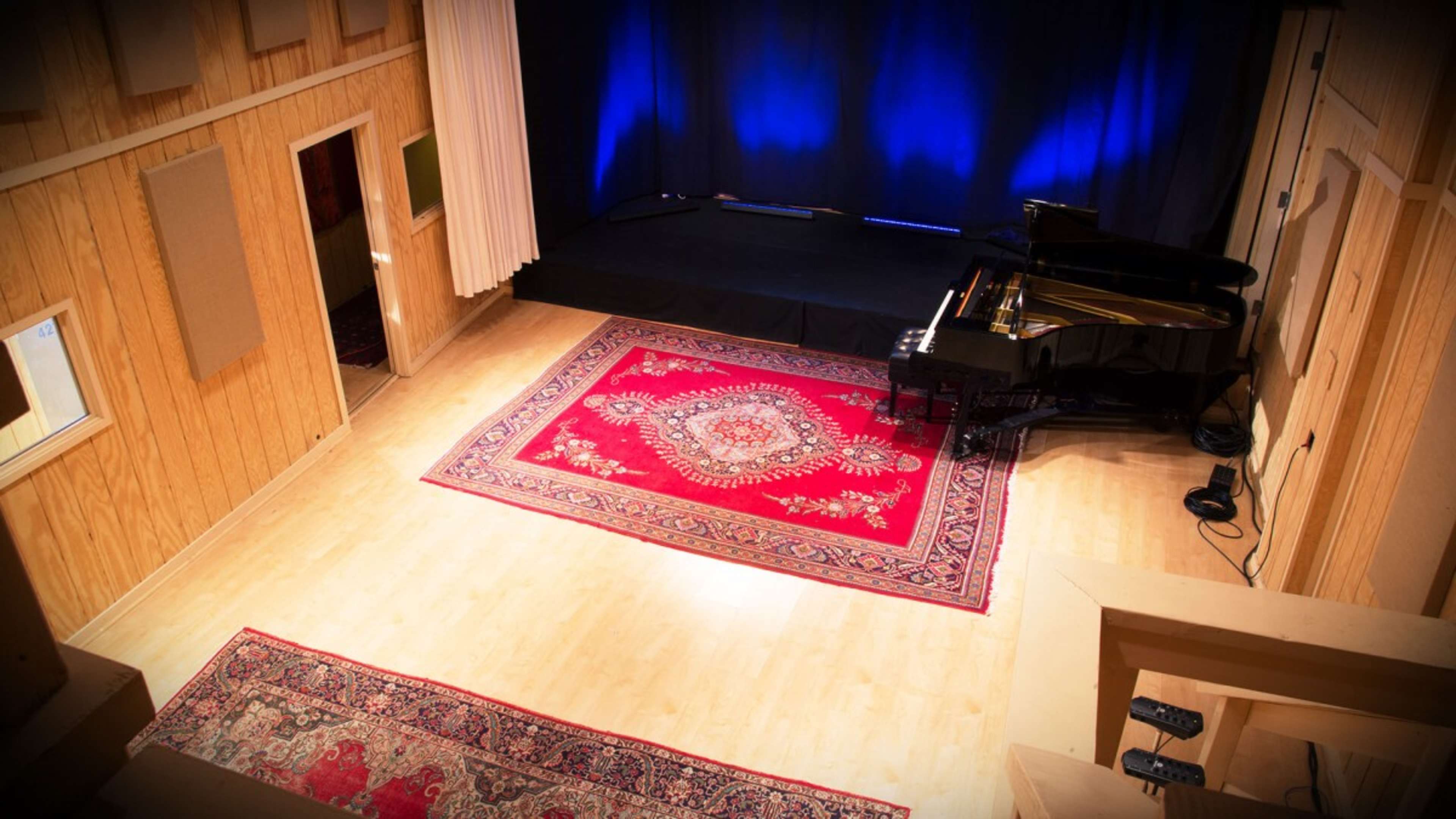 A small performance space with a grand piano on a red area rug, surrounded by wooden walls and blue lighting.