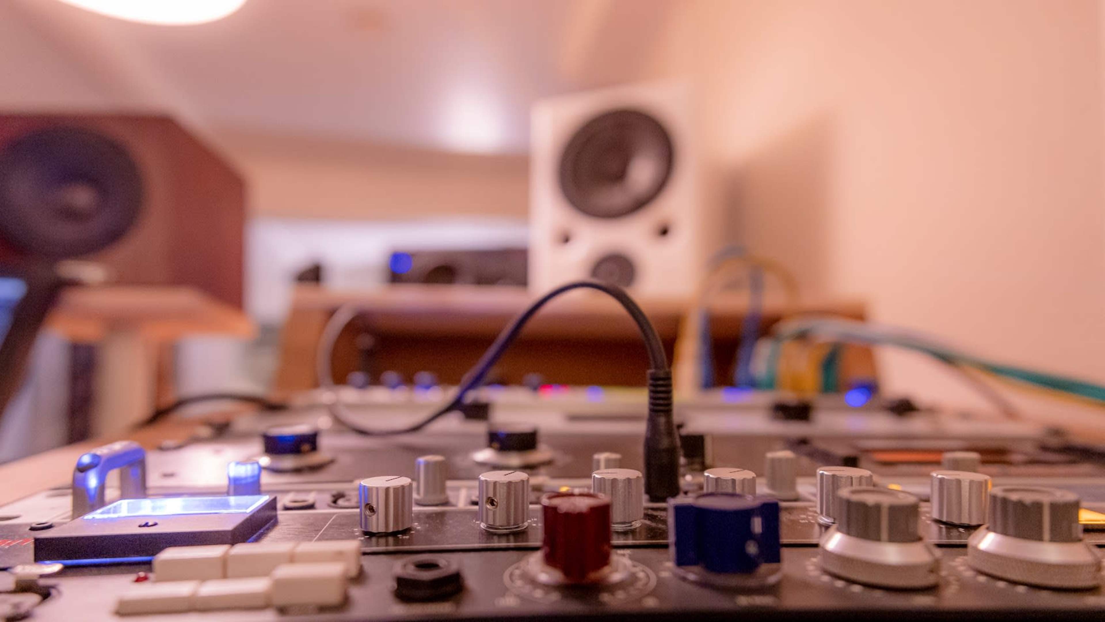 The image features a close-up view of a music mixing console with various knobs and connections, set against a blurred background of studio speakers and equipment.