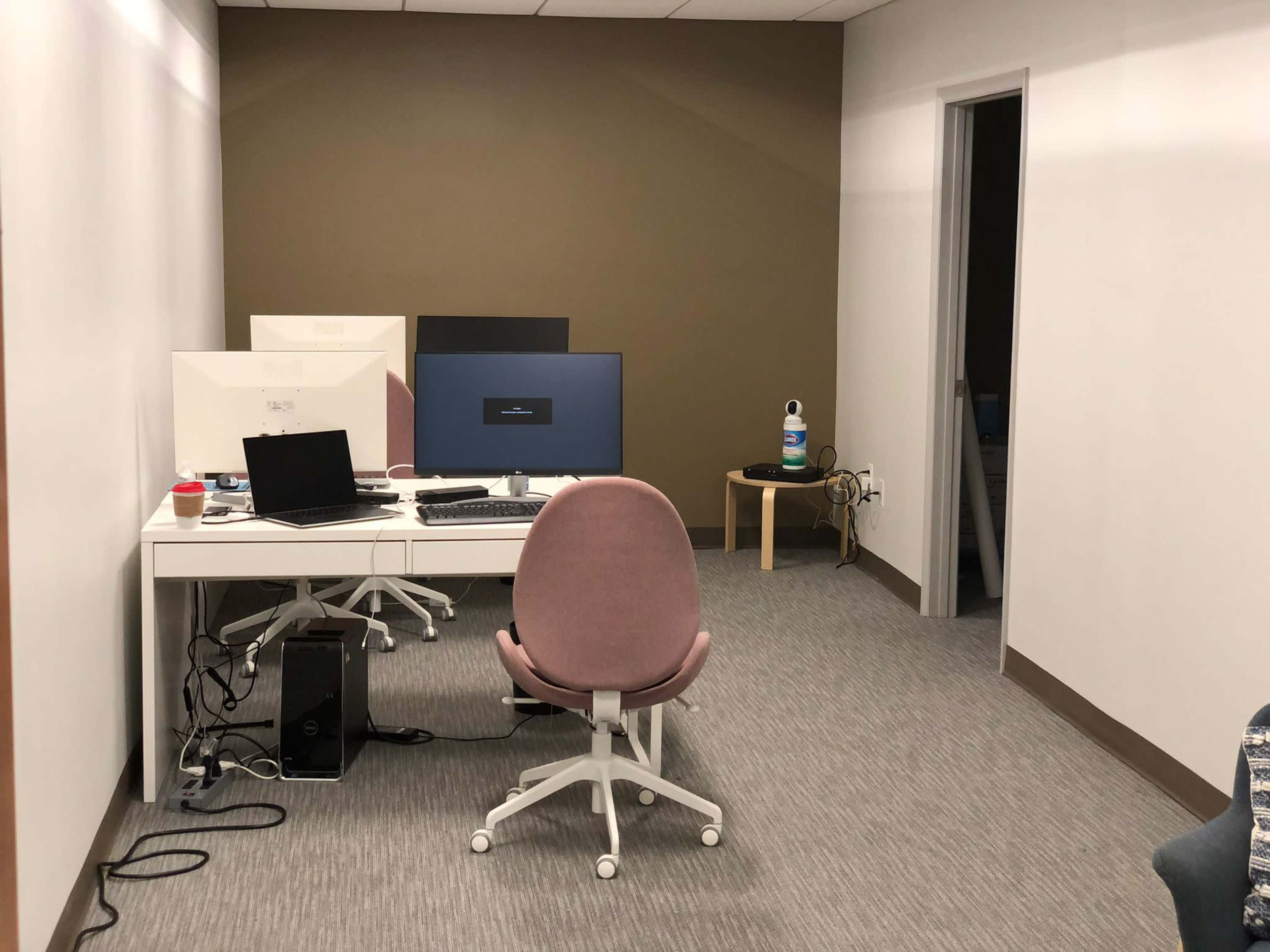The image shows a small office space featuring a desk with three computer monitors, a chair, and a water bottle on a small table.