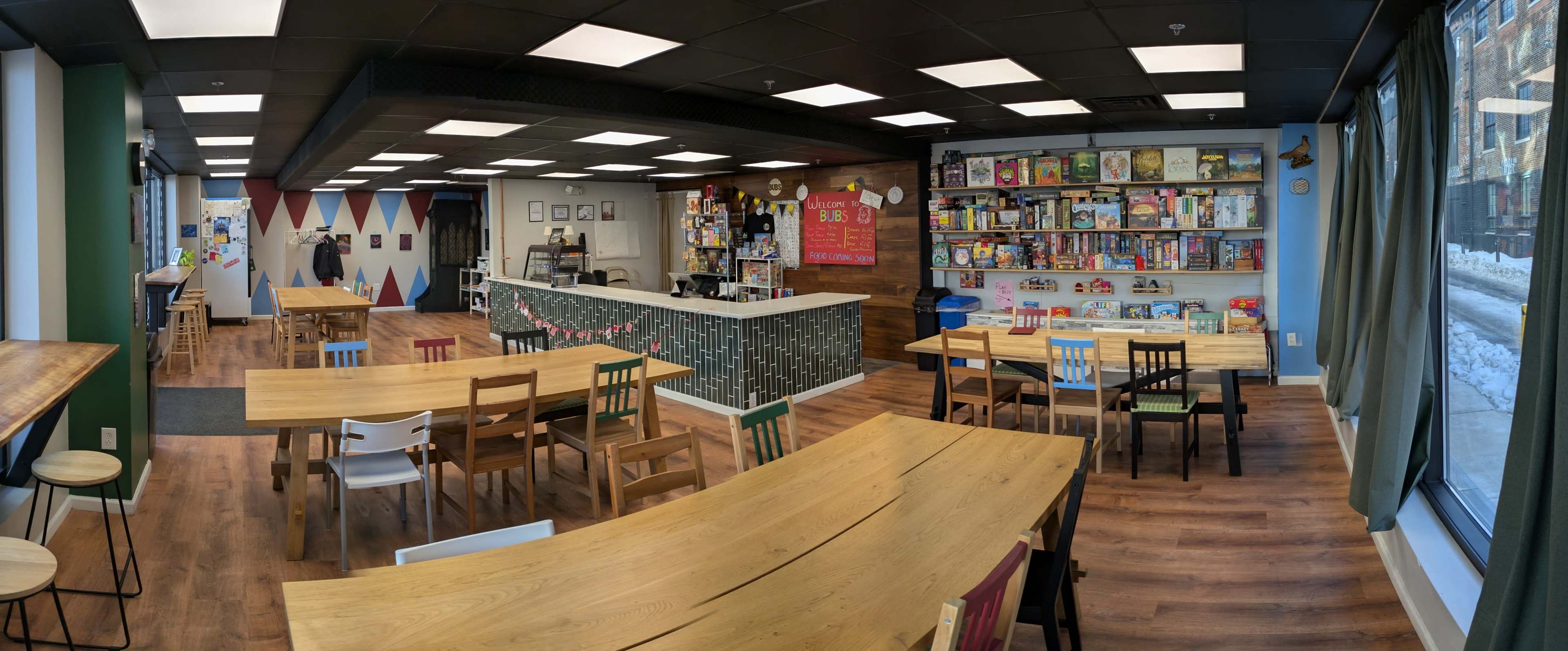 The image shows a cozy board game café featuring wooden tables, a counter with snacks, and shelves filled with various board games along one wall.
