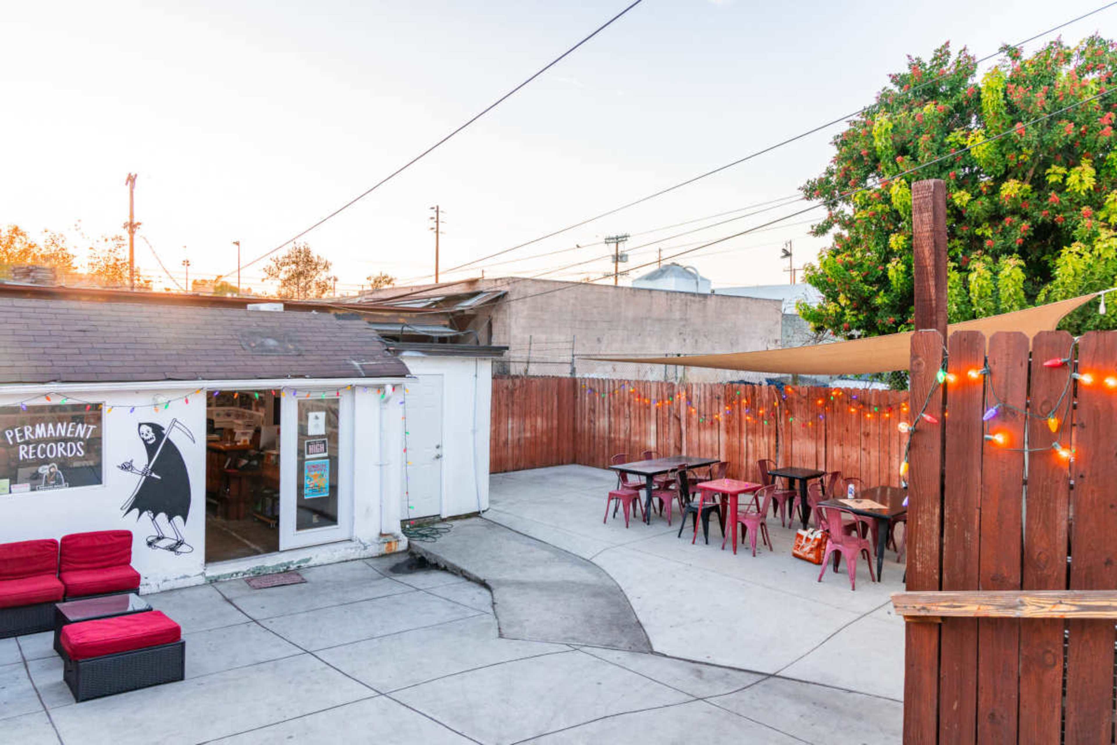 The image shows an outdoor seating area at a record store, featuring tables and chairs surrounded by a wooden fence and a tree, with string lights illuminating the space.