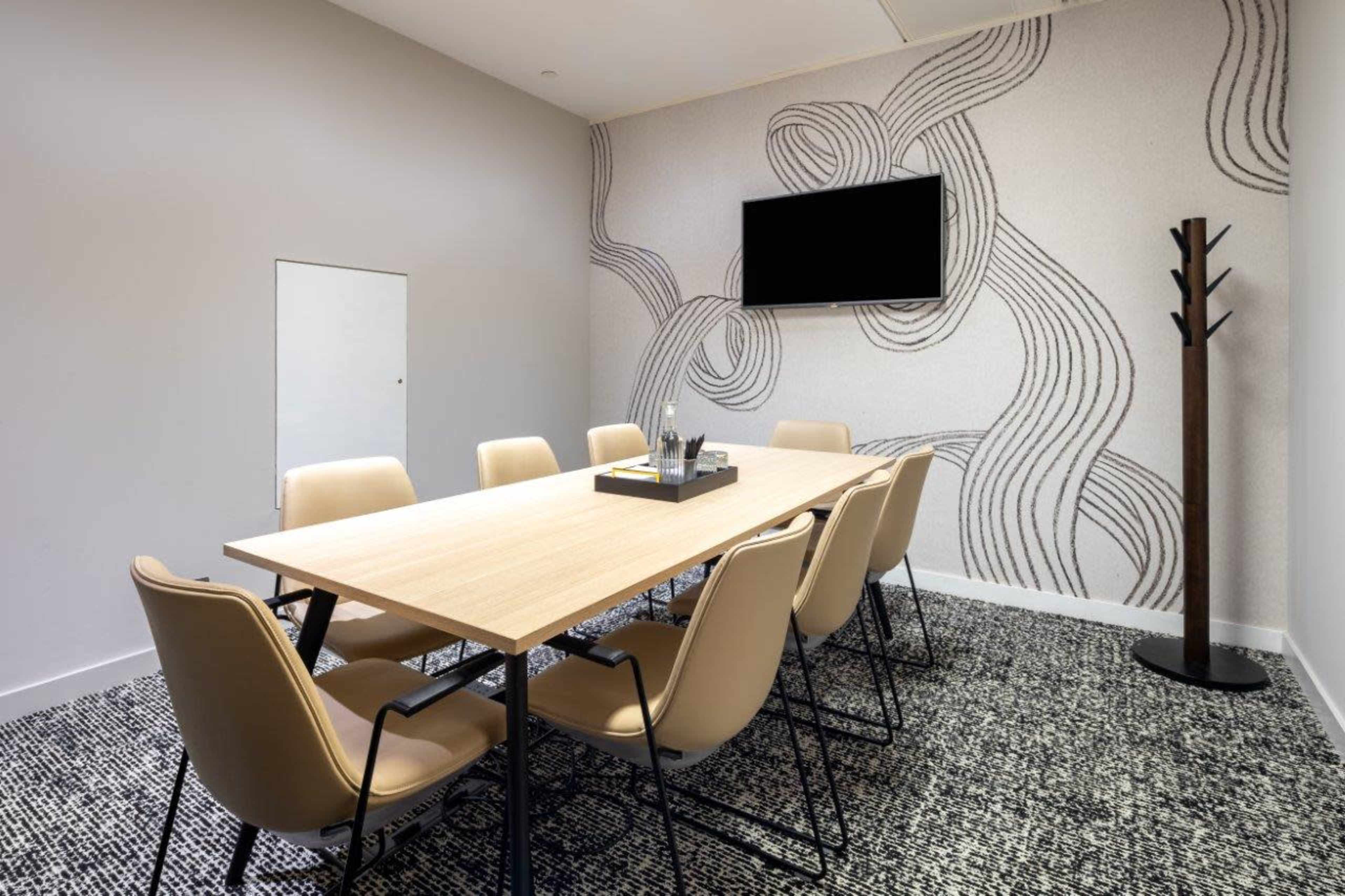 A modern conference room features a long wooden table surrounded by beige chairs, with a wall-mounted television and a decorative patterned wall.