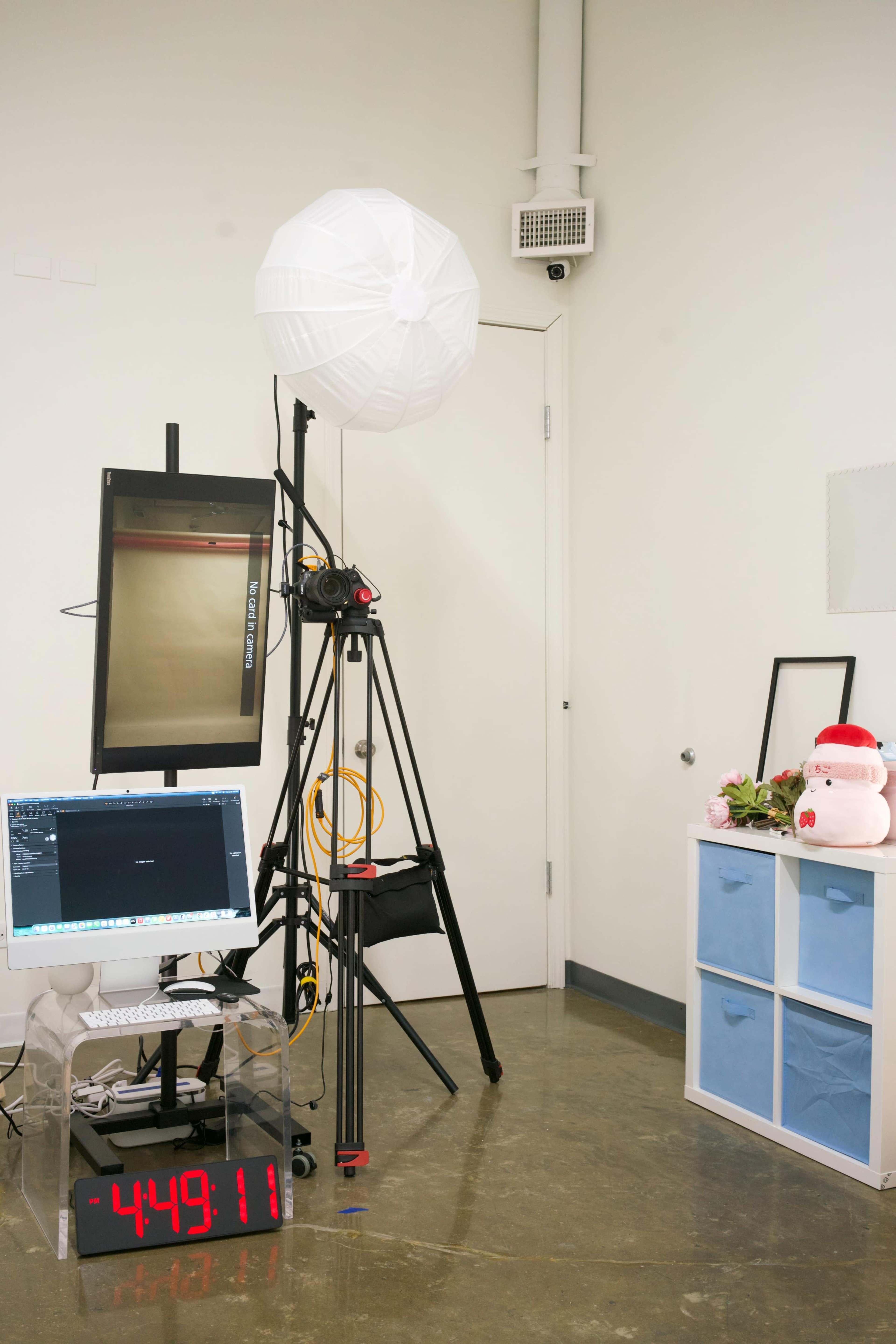 The image shows a photography setup in a studio, featuring a tripod-mounted camera, a light umbrella, a computer displaying software, and a large digital timer on the floor.