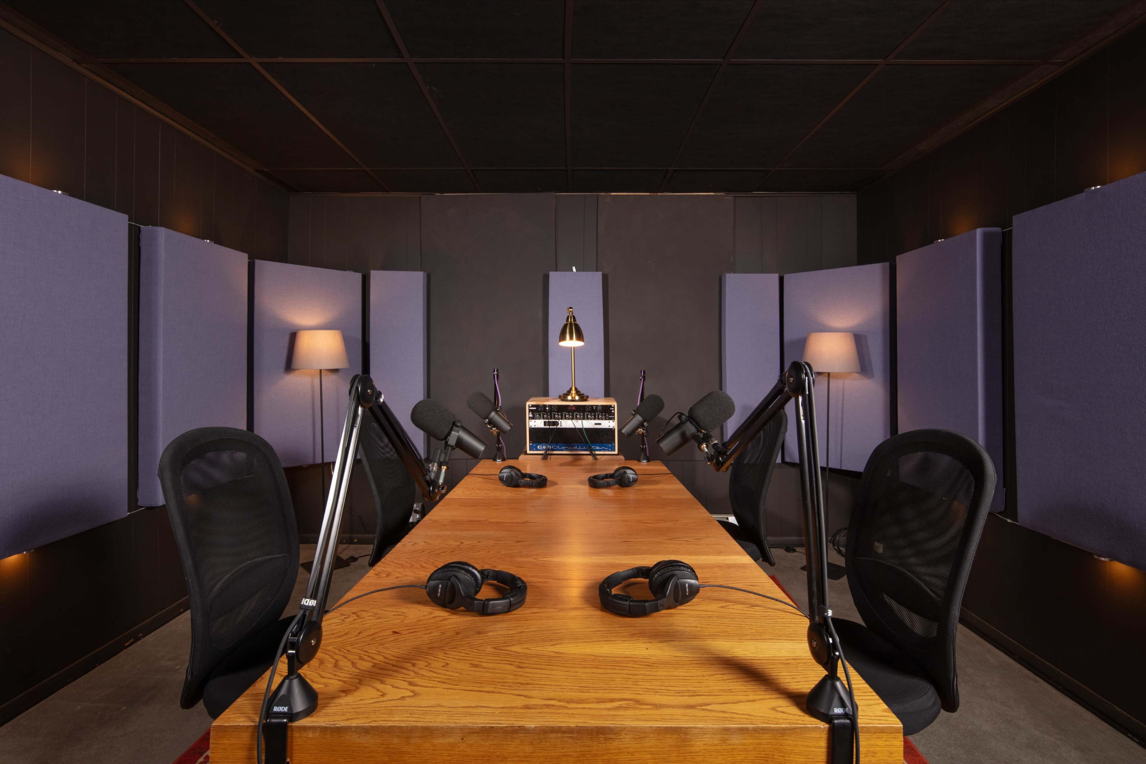 A recording studio features a large wooden table surrounded by equipped microphones, headphones, and soundproof wall panels.