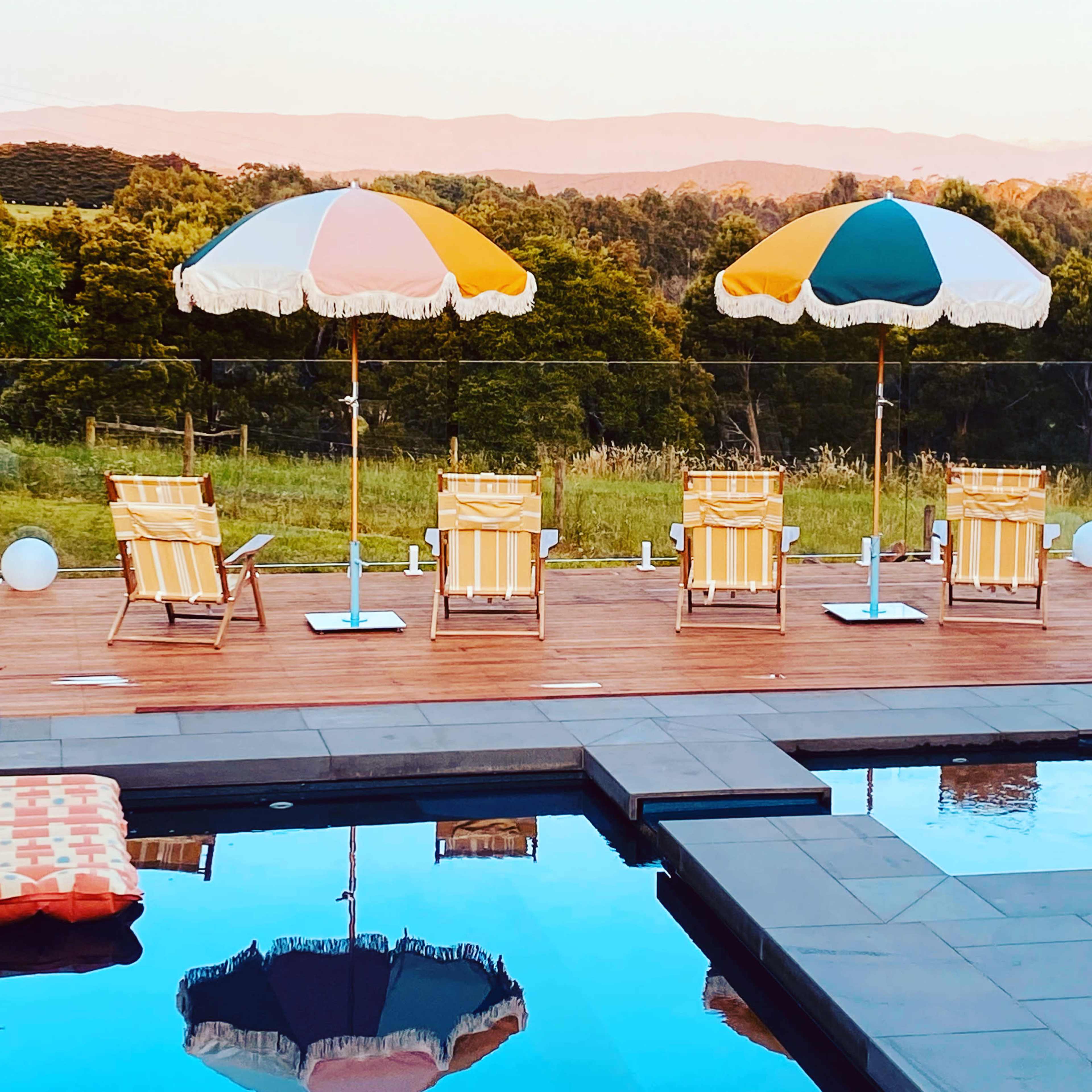 Four striped lounge chairs and umbrellas are arranged beside a pool reflecting greenery and mountains in the background.