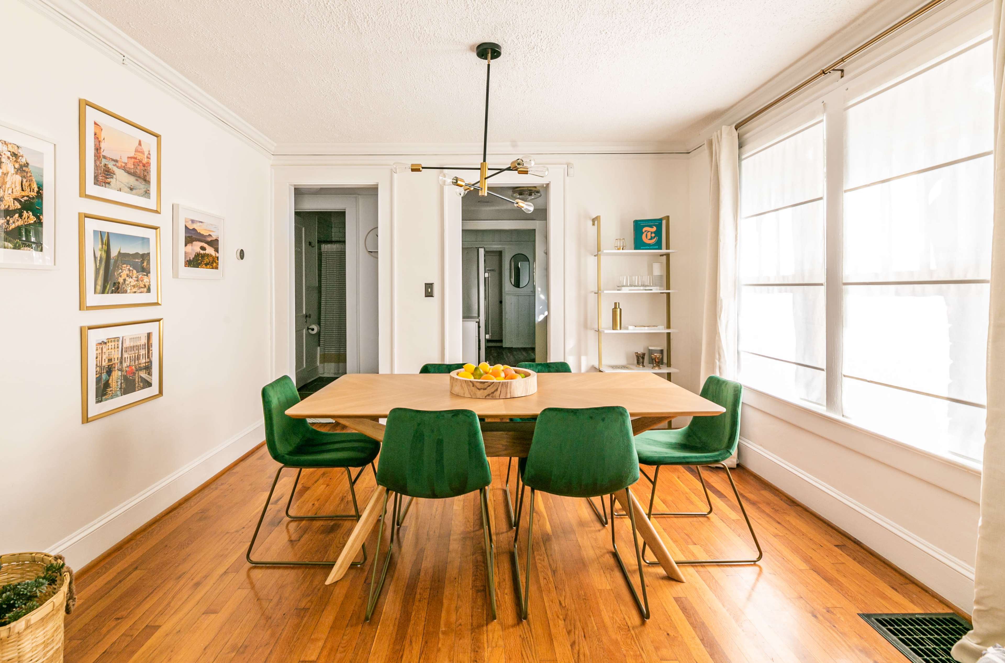 A dining room features a large wooden table surrounded by green chairs, with framed photographs on the walls and large windows letting in natural light.