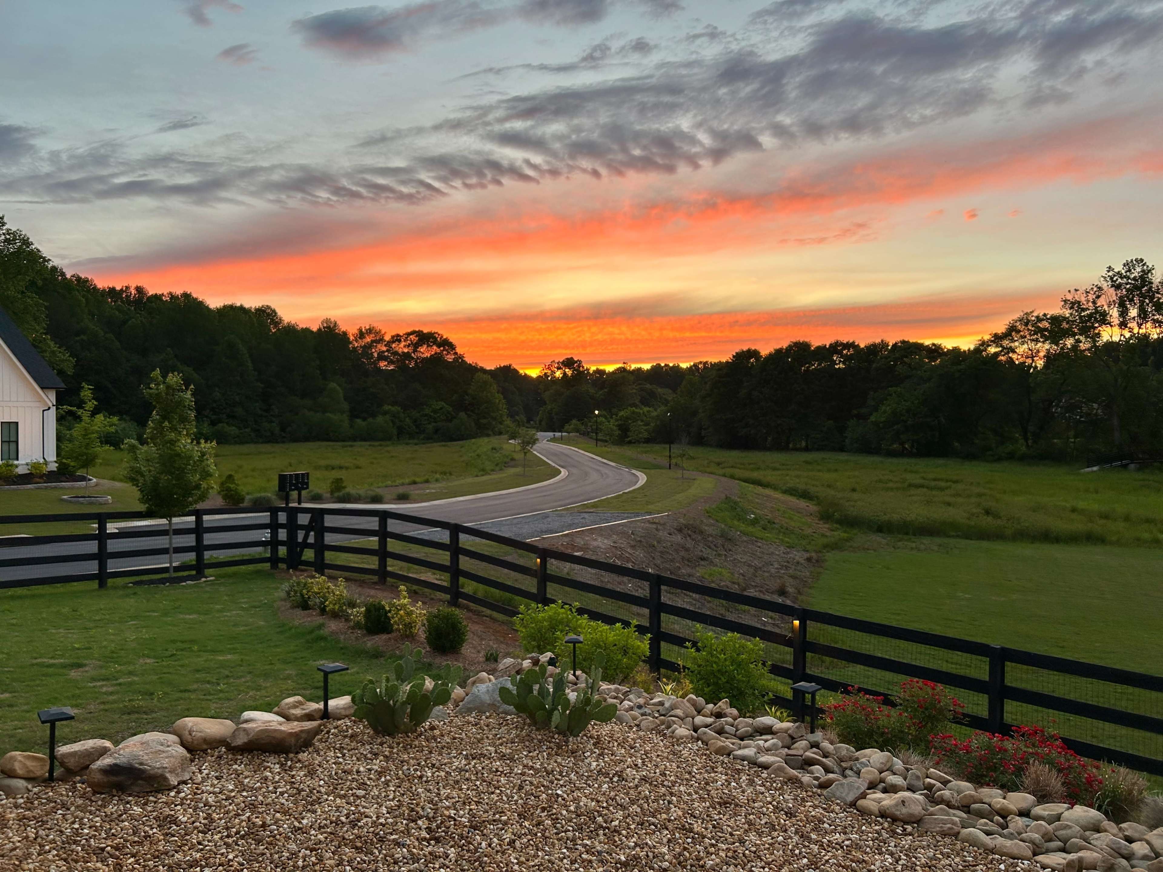 The scene shows a winding road bordered by a black fence, with a vibrant sunset casting colors across the sky and a landscaped foreground featuring rocks and plants.