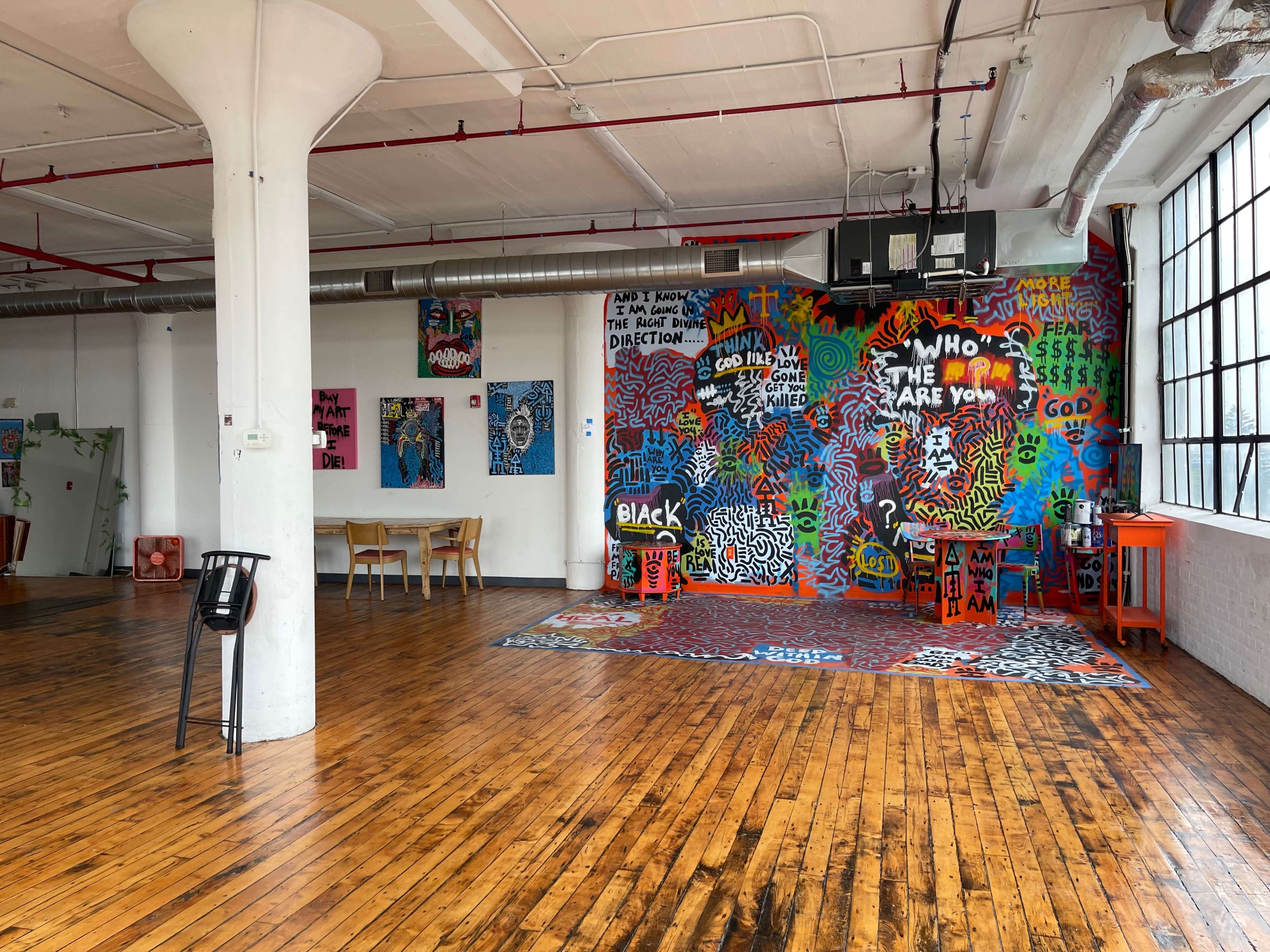 The image shows an industrial-style room with wooden floors, featuring a colorful graffiti-covered wall and several tables and chairs arranged in the space.