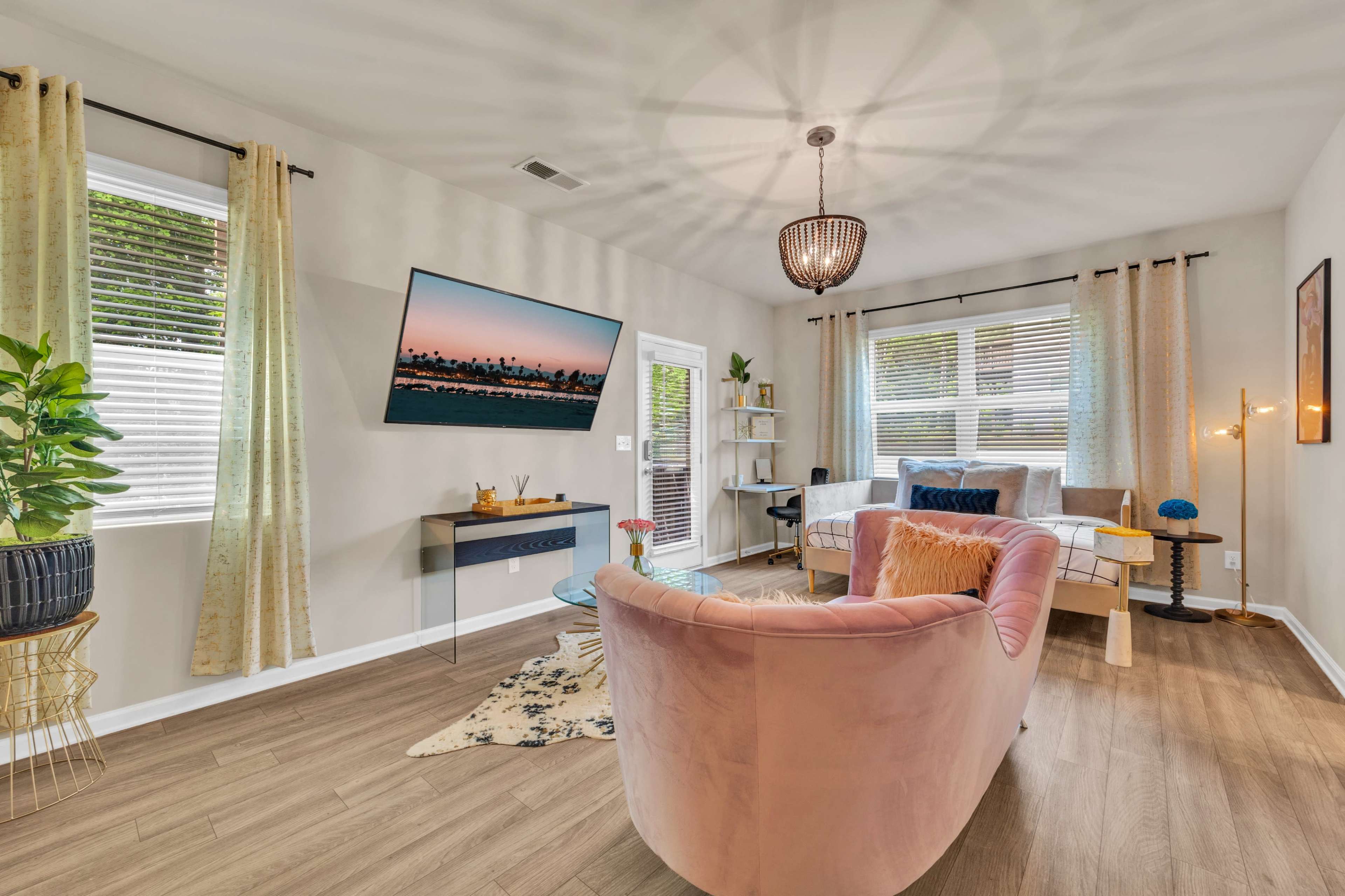 A brightly lit living room features a pink sofa, a television mounted on the wall, and large windows with light curtains.