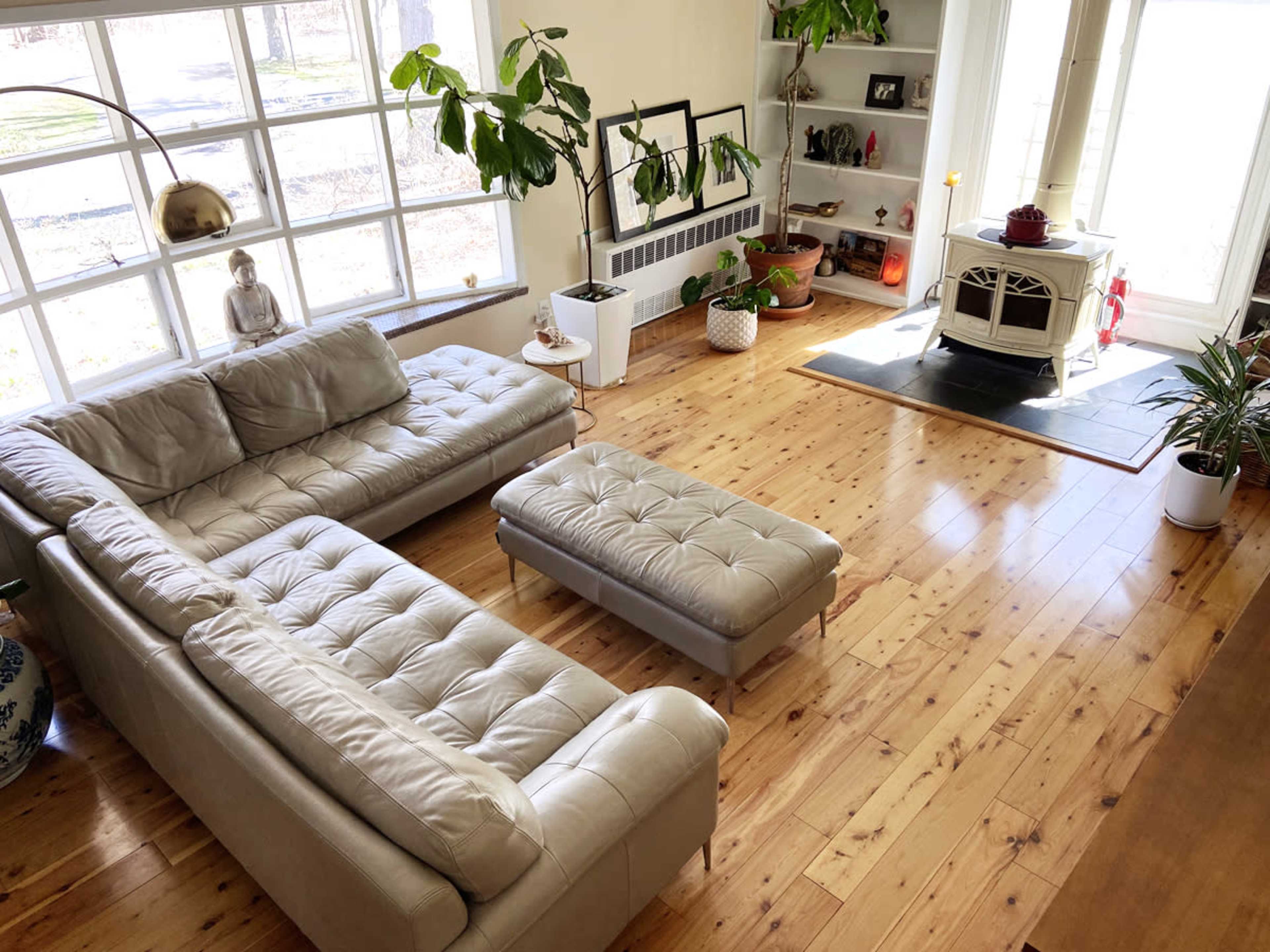 A light-colored leather sectional couch and ottoman are positioned in a sunlit living room with wooden flooring and large windows.