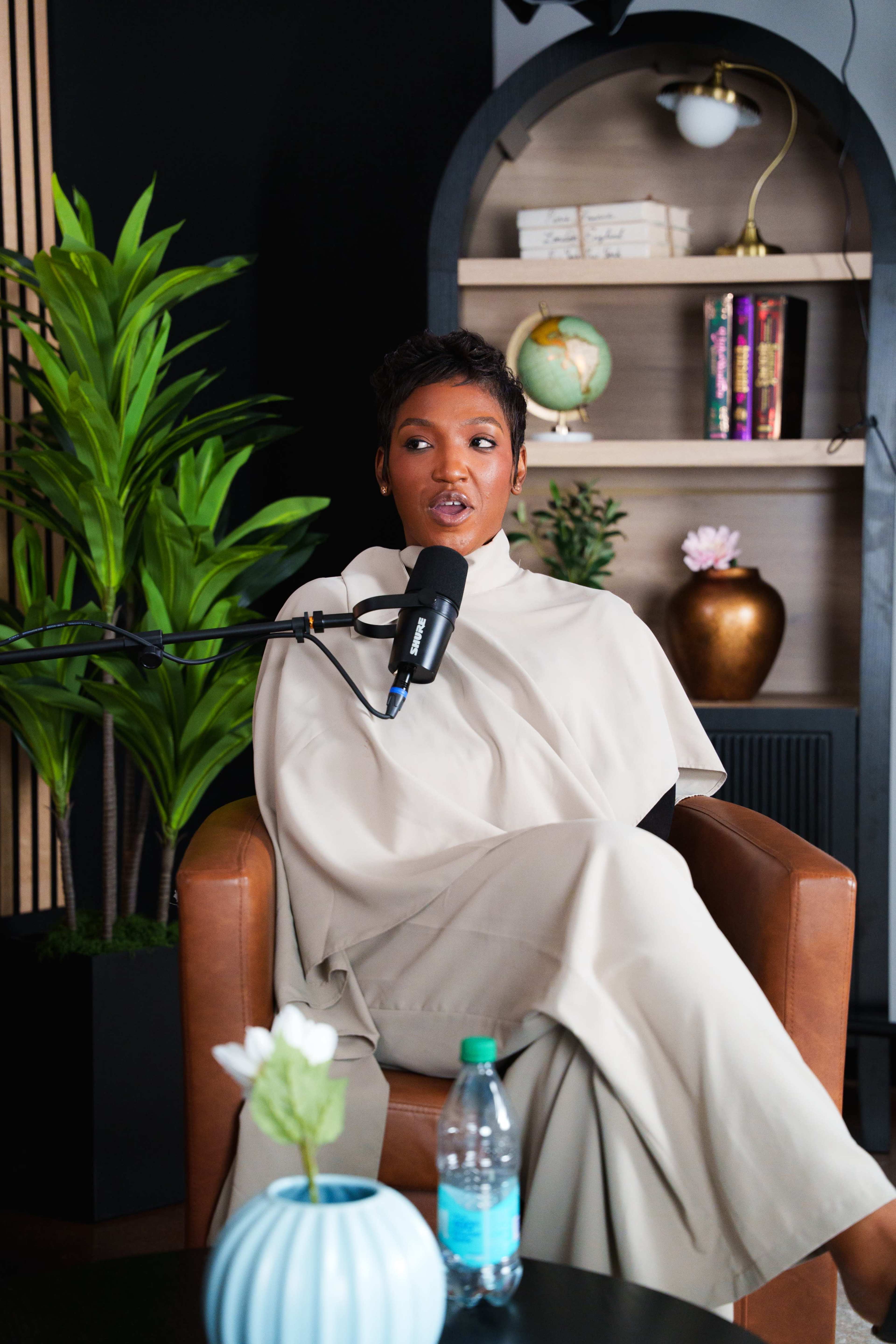 A woman dressed in a beige outfit sits in a brown leather chair with a microphone in front of her, surrounded by indoor plants and bookshelves.